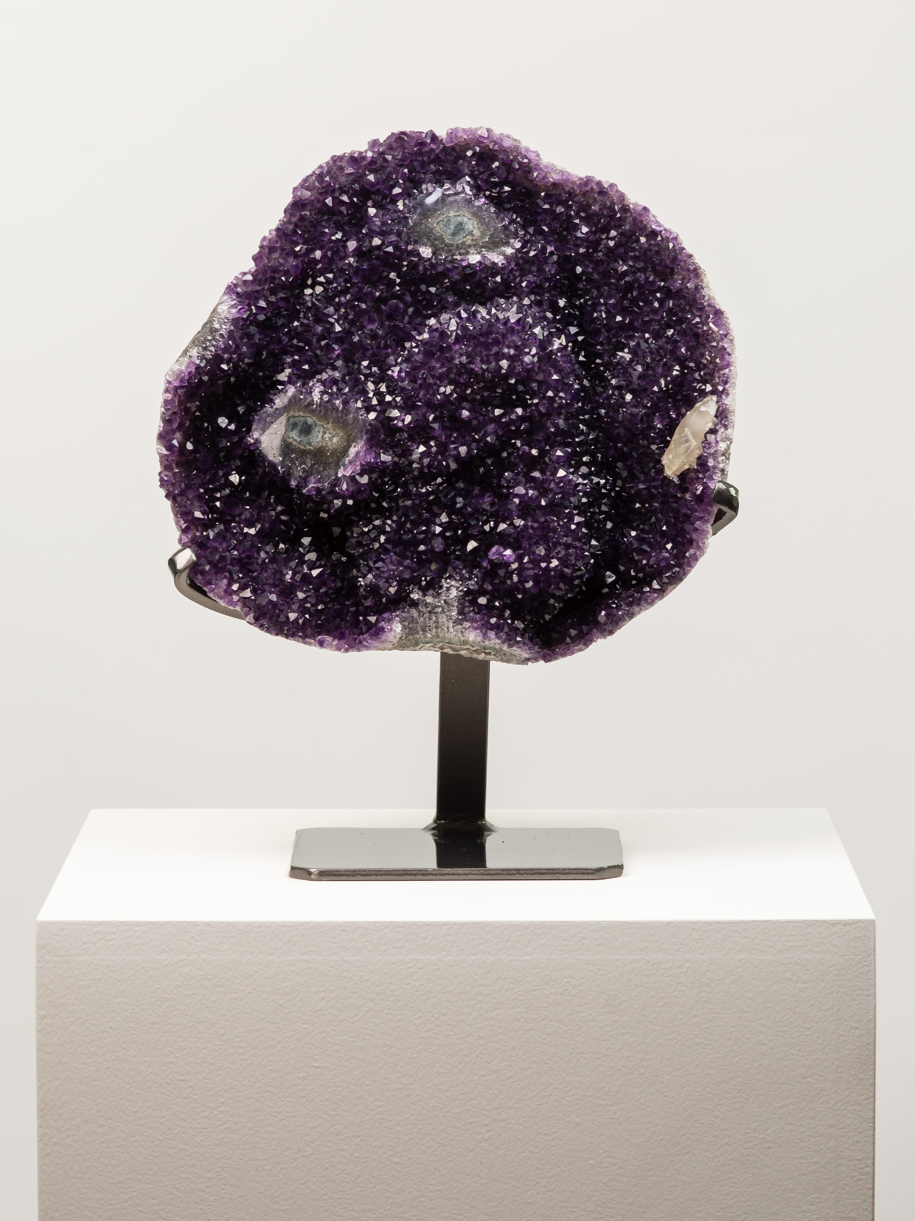 A beautiful oval shaped amethyst section with deep purple amethyst and two
cut amethyst eyes near the centre. A small calcite to the right completes the
composition.

This piece was legally and ethically sourced directly in the prestigious mines