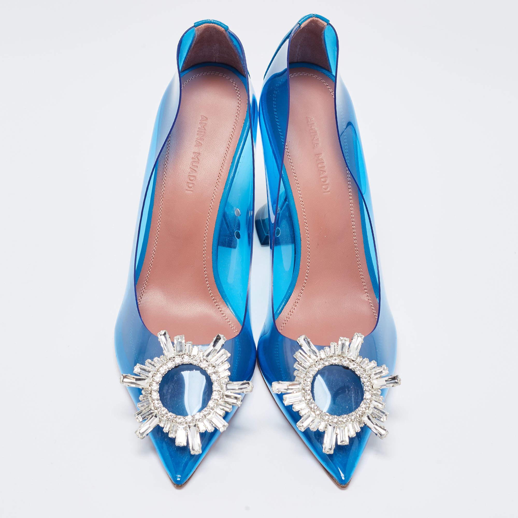 The Amina Muaddi Begum pumps are striking high-heeled shoes. They feature transparent blue PVC uppers, a pointed toe, and a sculpted heel with a signature flared design. These pumps exude modern elegance, combining transparency with a bold pop of