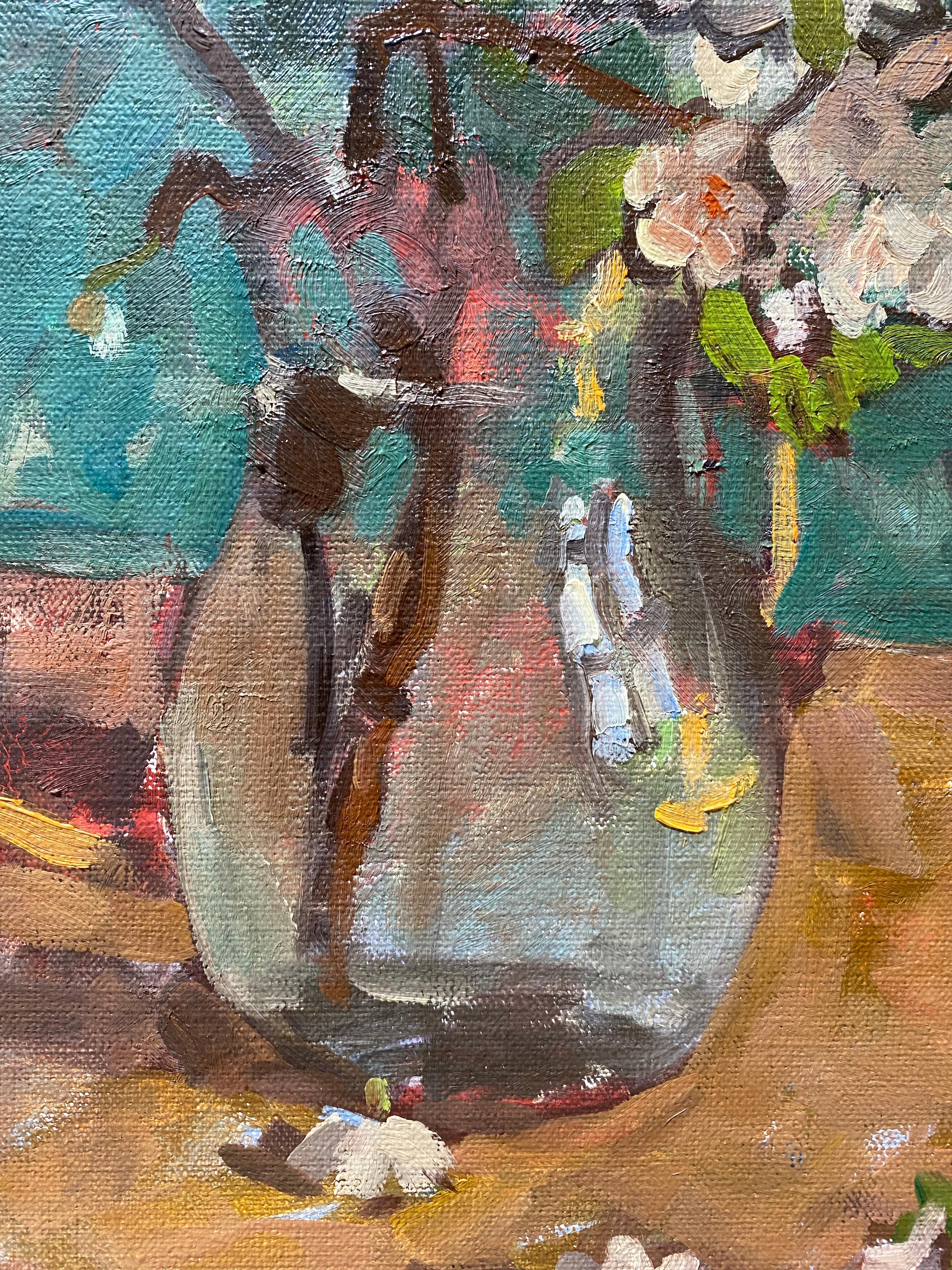 A still life oil painting of branches of pear blossoms, freshly cut and arranged in a glass pitcher of water. These spring blossoms flourish in small white petals, against curved brown branches. The flowers are set atop a wooden table, and in front