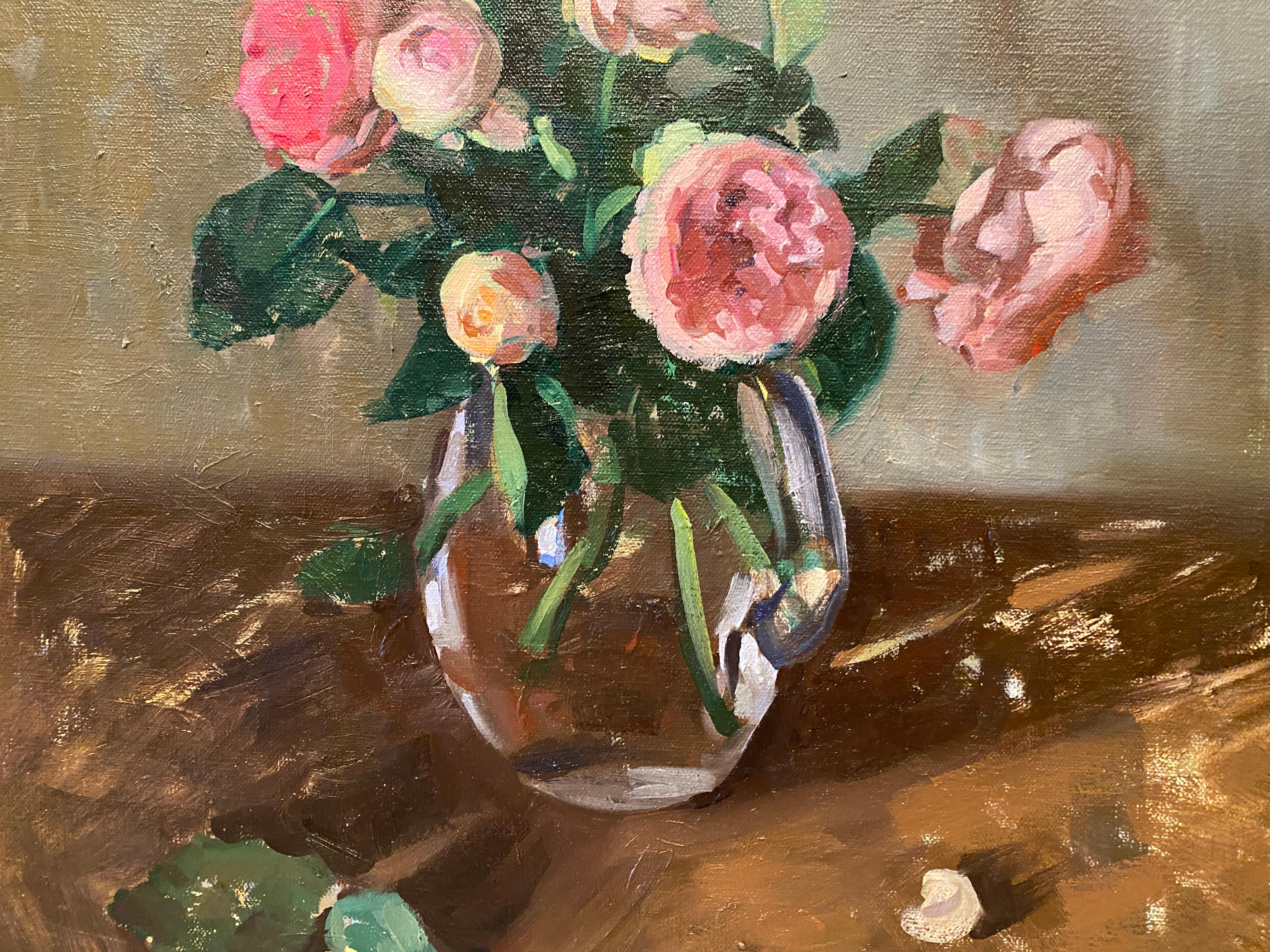 An elegant still-life painting of pink roses in a round glass pitcher atop a table. Placed atop a natural wood tabletop and against a pale grey wall, the flowers pop out with color and light. 

Painting Dimensions: 23.62 in x 19.68 in
Framed