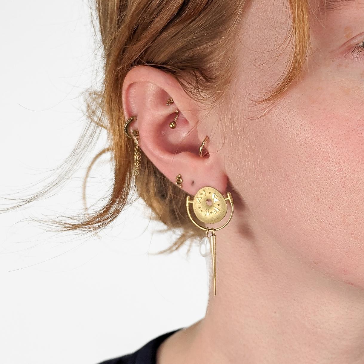 A fine pair of Amy Roper Lyons 18k gold earrings.

In 18k yellow gold. 

By Amy Roper Lyons.

With a donut shape in center and a half circle bar that supports two dangling pendant charms  - a freshwater pearl and a spear shape.  

Amy Roper Lyons is