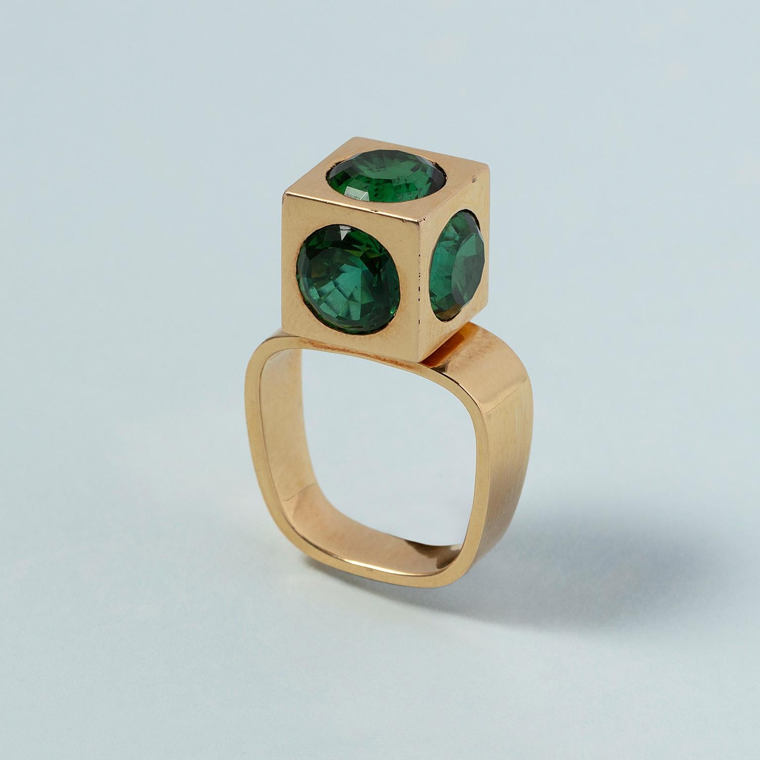 An 18 carat yellow gold ring with a gold cube on top set with five facetted round-step cut bright green tourmalines, (bi-color, total: 11.46 ct), the shank is a rounded square, signed: Trudel, for Rudolf Trudel, Switzerland, circa 1970. 

weight:
