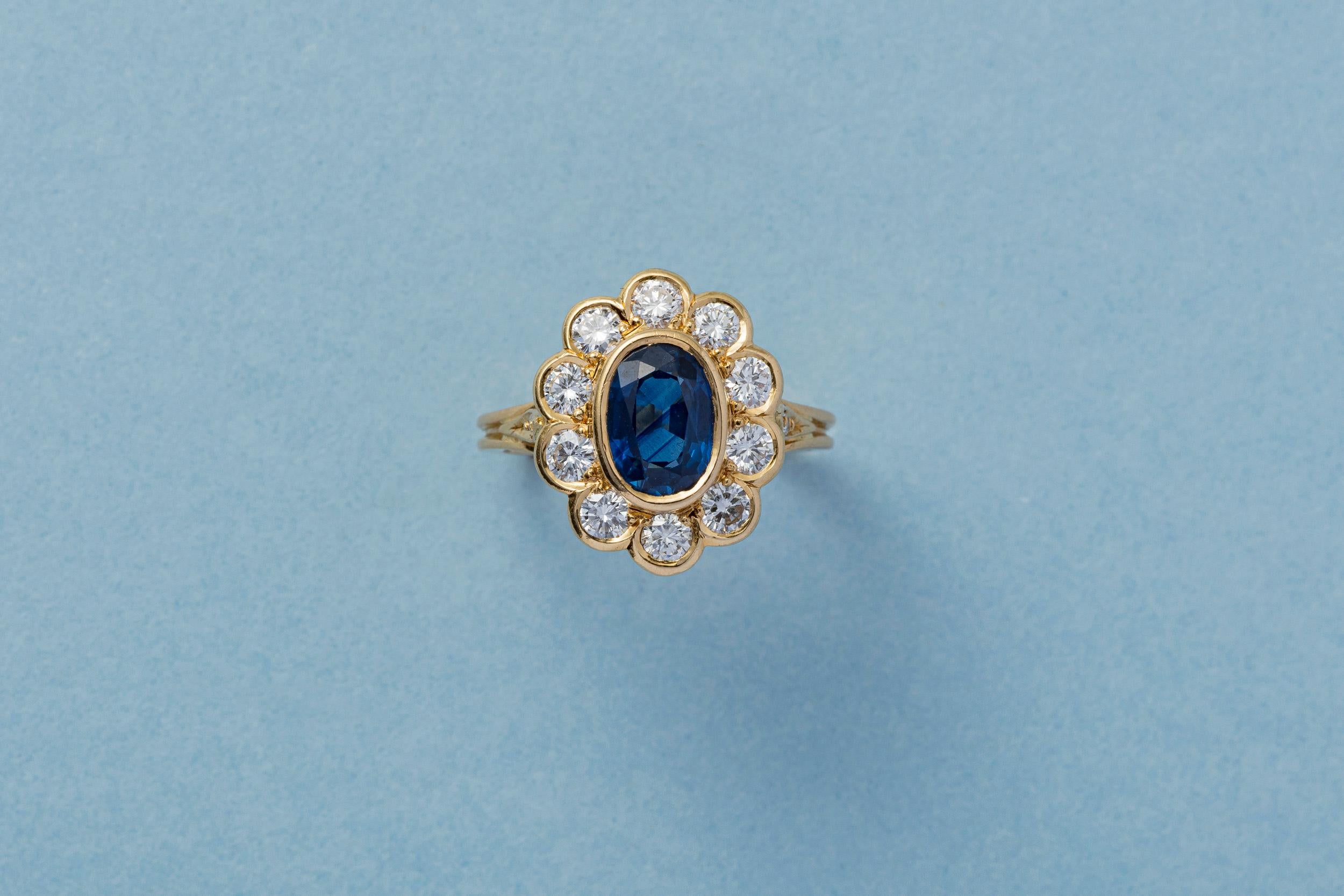 An 18 carat yellow gold cluster ring set with an oval, facetted heated sapphire (app. 1.5 carat, with NEL certificate) surrounded by ten brilliant cut diamonds. All in closed settings with the most gorgeous gallery with open hearts and with on the