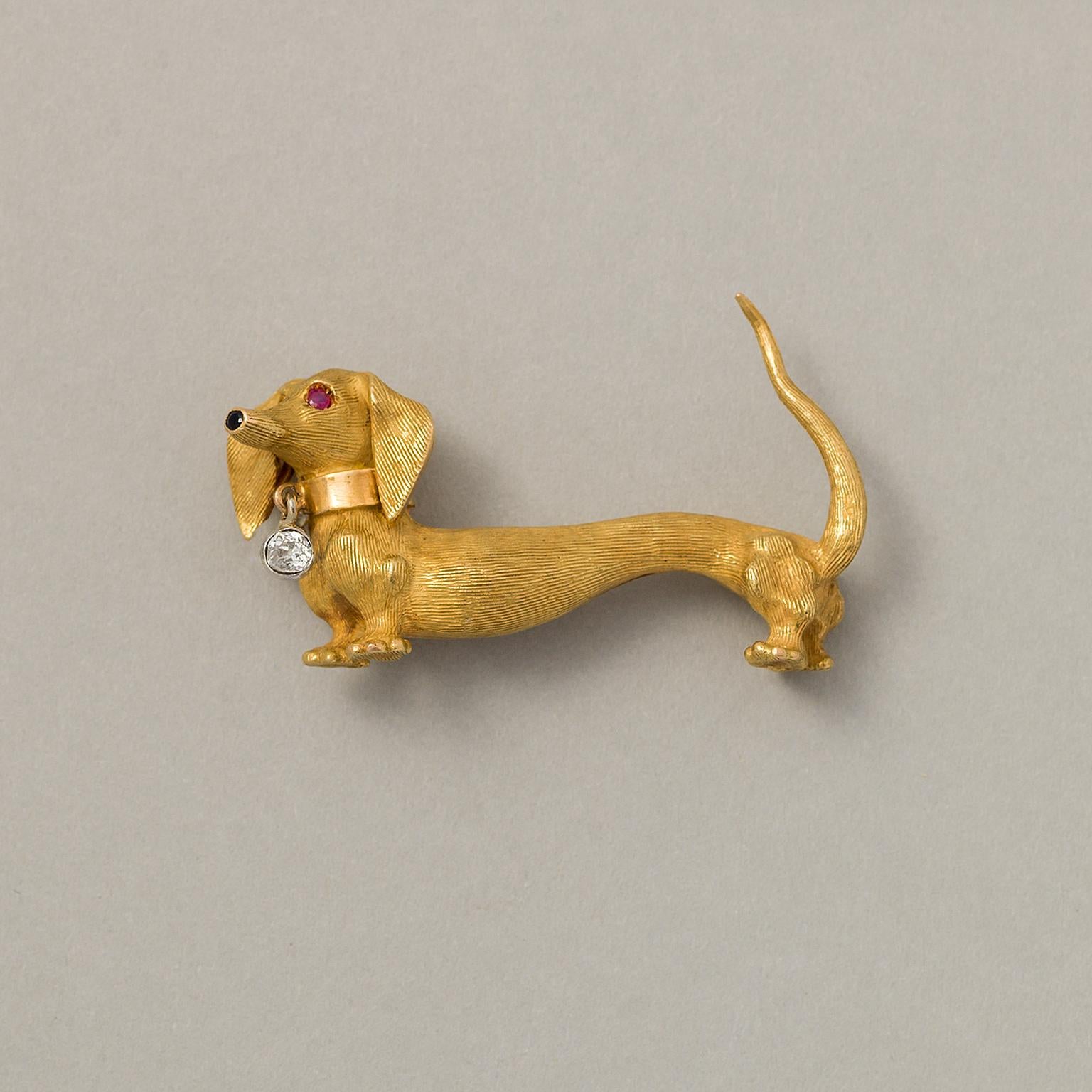 An 18 carat gold happy little Dachshund brooch with a matte striped fur and a polished collar with a dangling old cut diamond set in white gold with an onyx nose and ruby eyes, French, circa 1960.

weight: 14.78 grams
dimensions: 4.4 x 2.5 cm