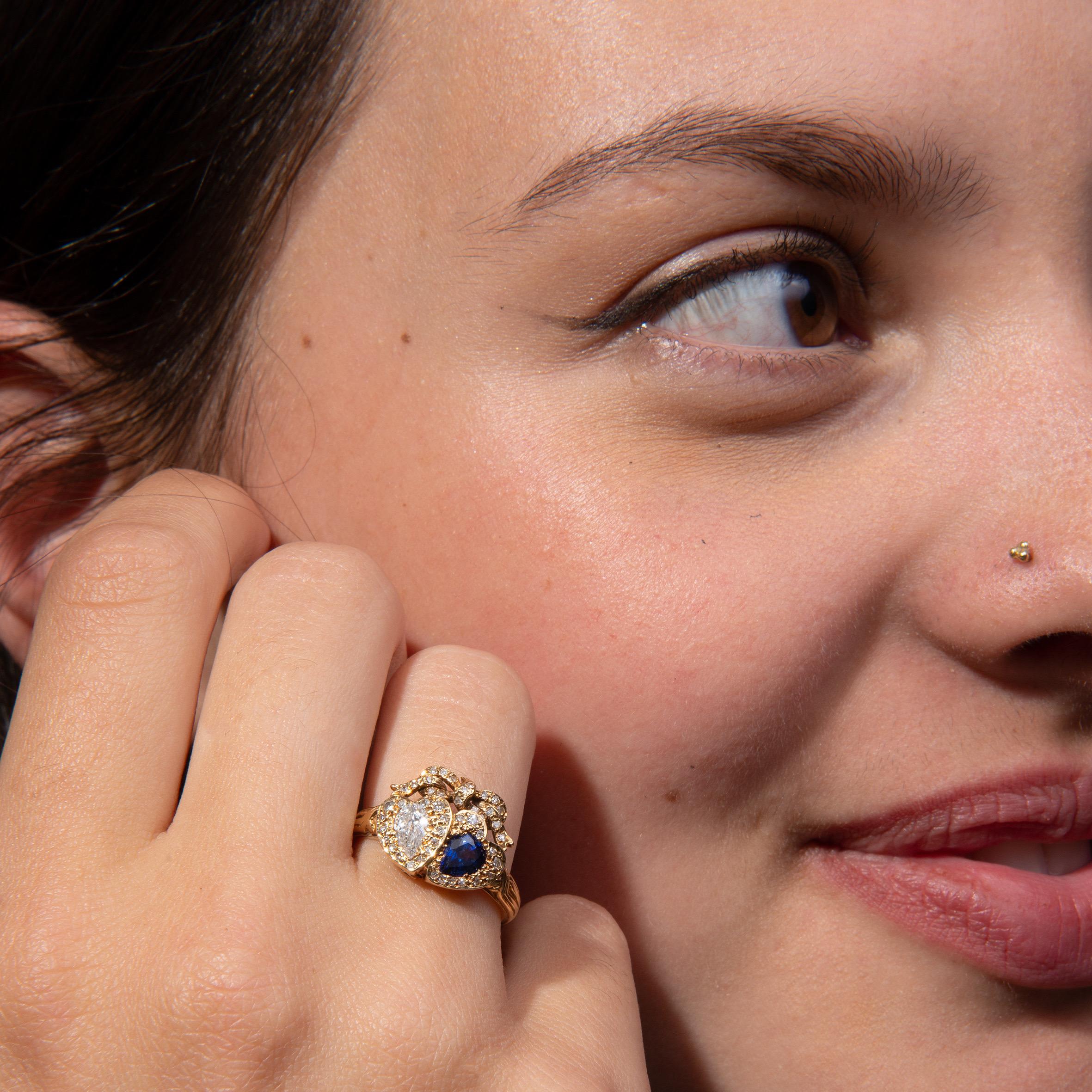 A pretty 18 carat yellow gold double heart ring with a pear-shaped faceted diamond (app. 0.30 carat) and a pear shaped sapphire, set in a diamond-encrusted heart-shaped setting (app. 0.50 carat in total). Crowned by a bow inlaid with small diamonds.