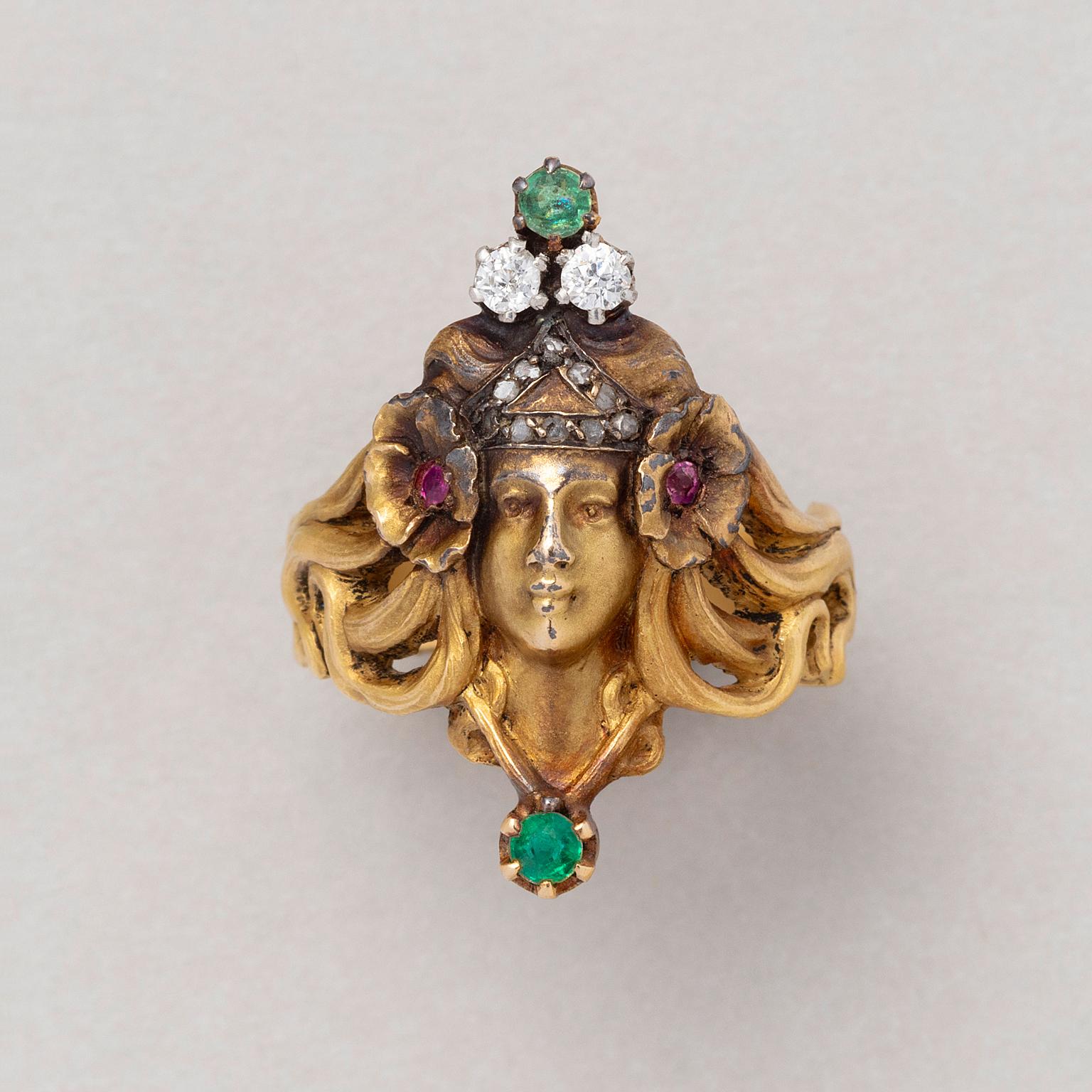 An 18 carat gold Art Nouveau ring featuring a Nymph with long waving hair wearing a tiara set with ten rose cut diamond and on top with two old cut diamonds and an emerald in her hair are two flowers with rubies in the heart of the flowers, France,
