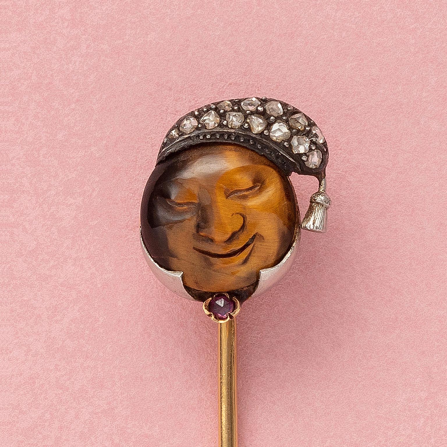 An 18 carat yellow gold stick pin with a round tiger eye engraved with a the face of a sleeping moon. He wears a silver night cap set with small rose cut diamonds and with a tassel. Under its chin a small rose cut ruby set in a flower shape gold