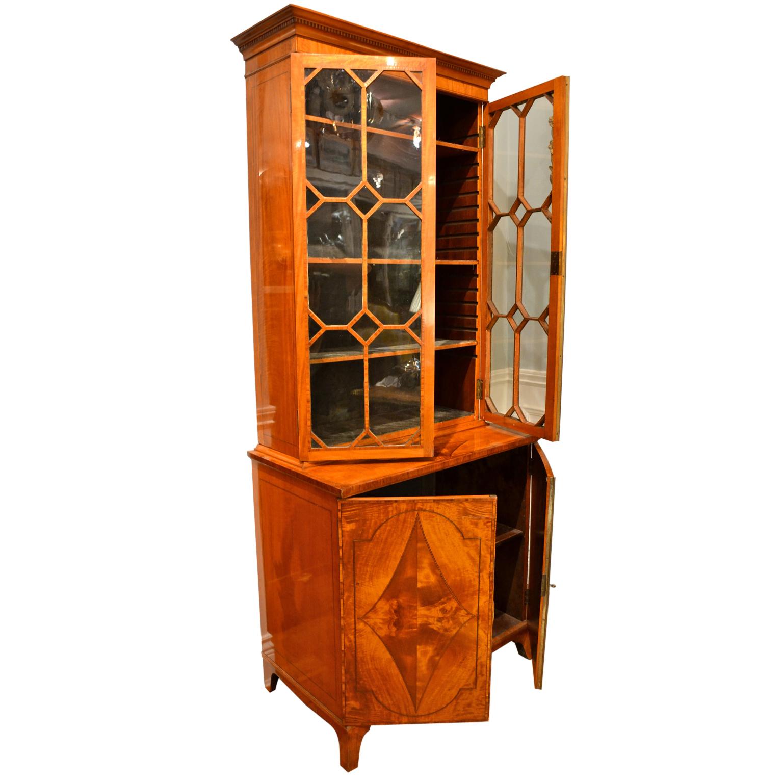 A classic period Sheraton English satinwood and tulipwood China display case or bookcase the upper section with dentil cornice above a pair of astragal glazed doors, enclosing three adjustable shelves, the base consisting of a shelved cupboard
