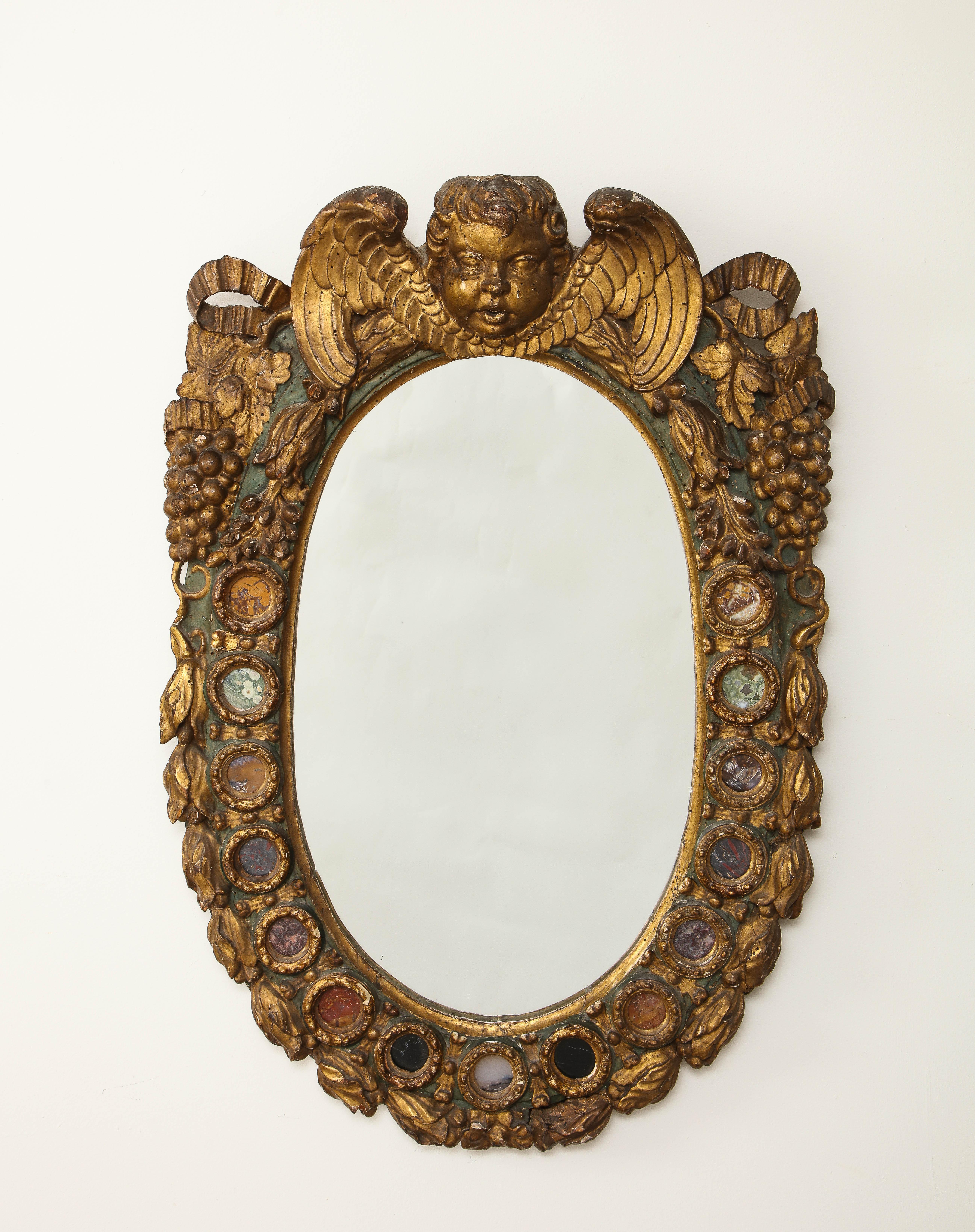 Un incredibile e raro specchio barocco italiano del XVIII secolo intagliato e in legno dorato con pietre dure intarsiate. La cornice di forma ovale è riccamente ornata da una maschera di cherubino alata sullo stemma. Il bordo è decorato con motivi
