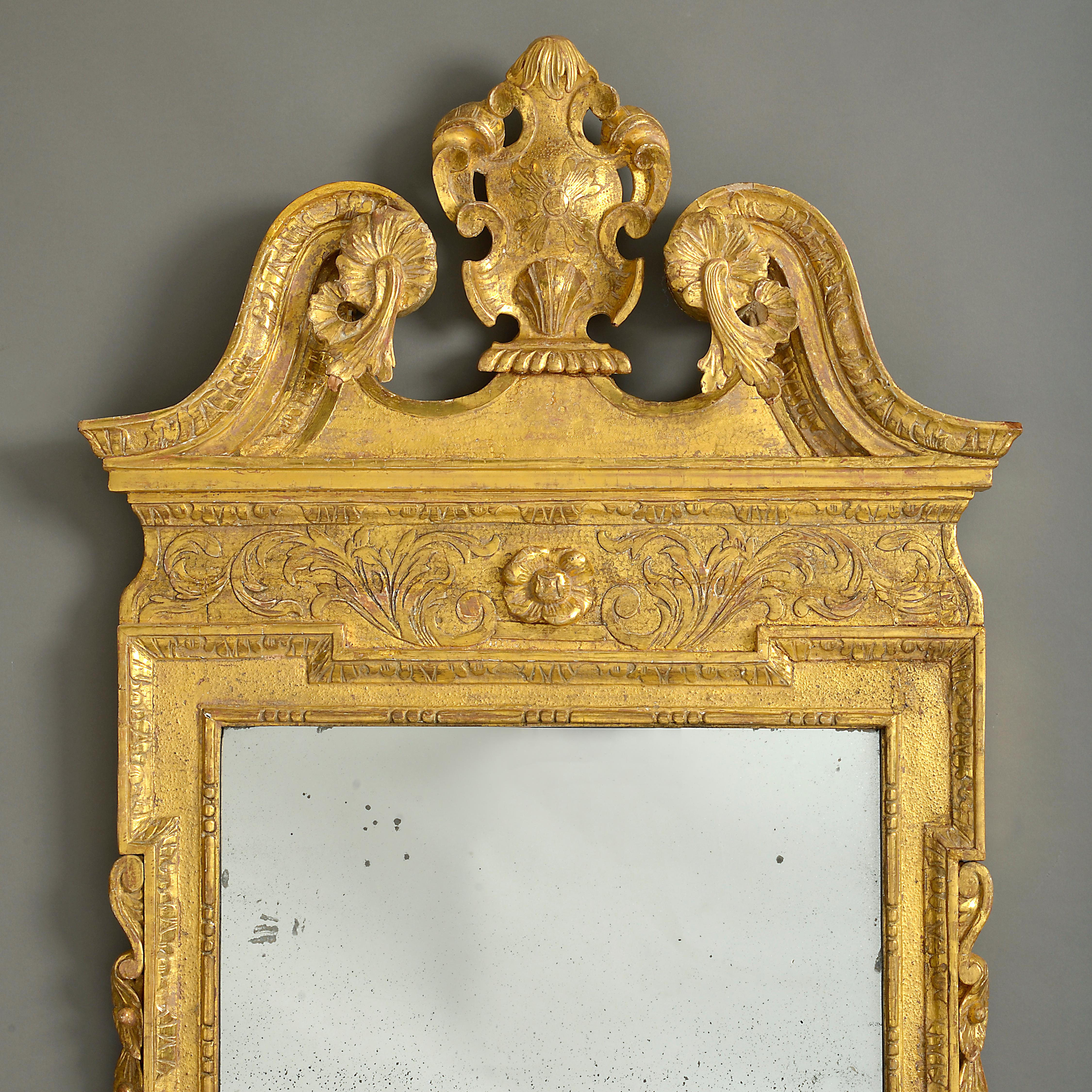 An early 18th century George II Period giltwood mirror in the manner of William Kent, the broken swan-neck pediment centred by a cartouche above a mercury glass plate within a panelled and sanded surround, flanked by trailing festoons of leaves and