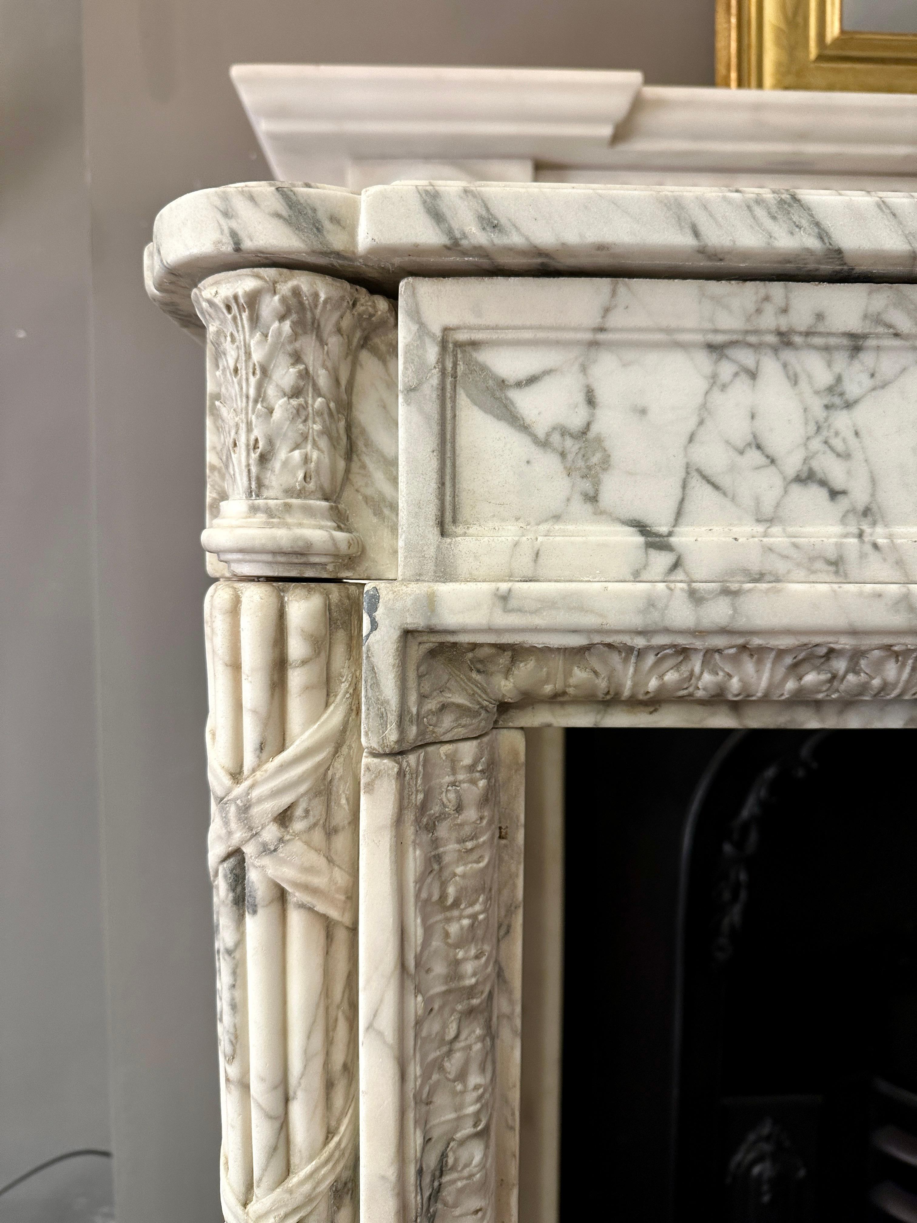An 18th century period Louis XVI fireplace executed in Italian Calacatta marble. Compact and attractive with slim tapering ribbon crossed fluted columns with Acanthus leaf corner blocks. The frieze with simple plain fielded panel to front. The