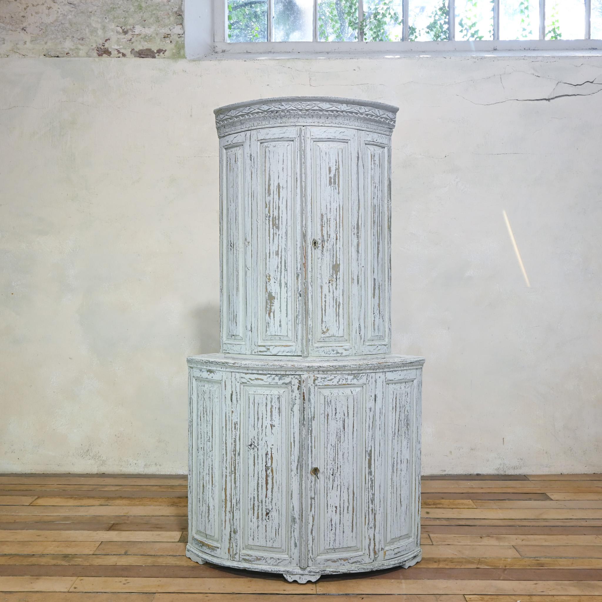 An 18th-century Swedish Gustavian corner cupboard, featuring an original dry-scraped finish that reveals a delicate patina with subtle tones of white and light grey. The elegant design includes a gracefully rounded top with ornate carved detailing,