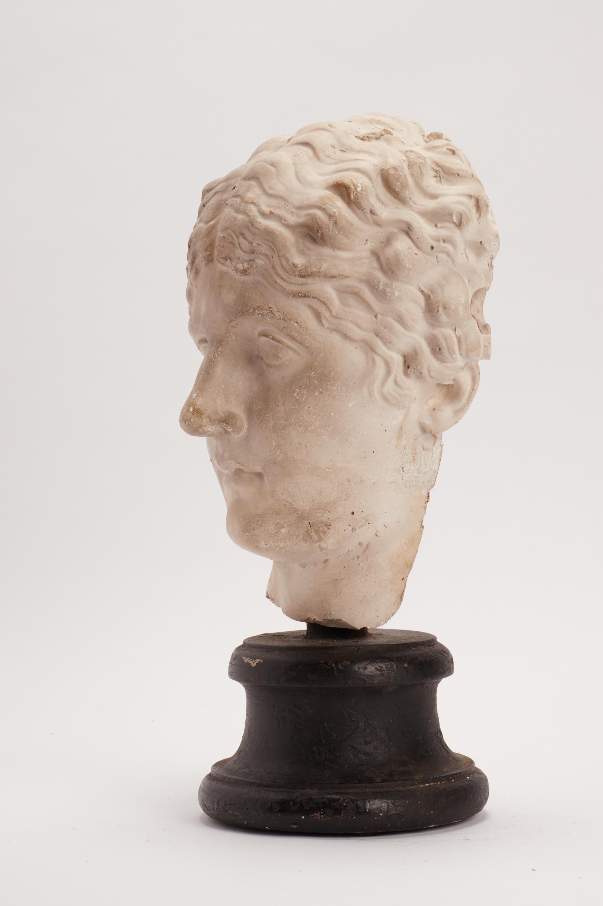 Over the wooden black painted base is set the plaster cast of a roman women’s head. The cast for drawing teaching in academy, Italy, circa 1890.