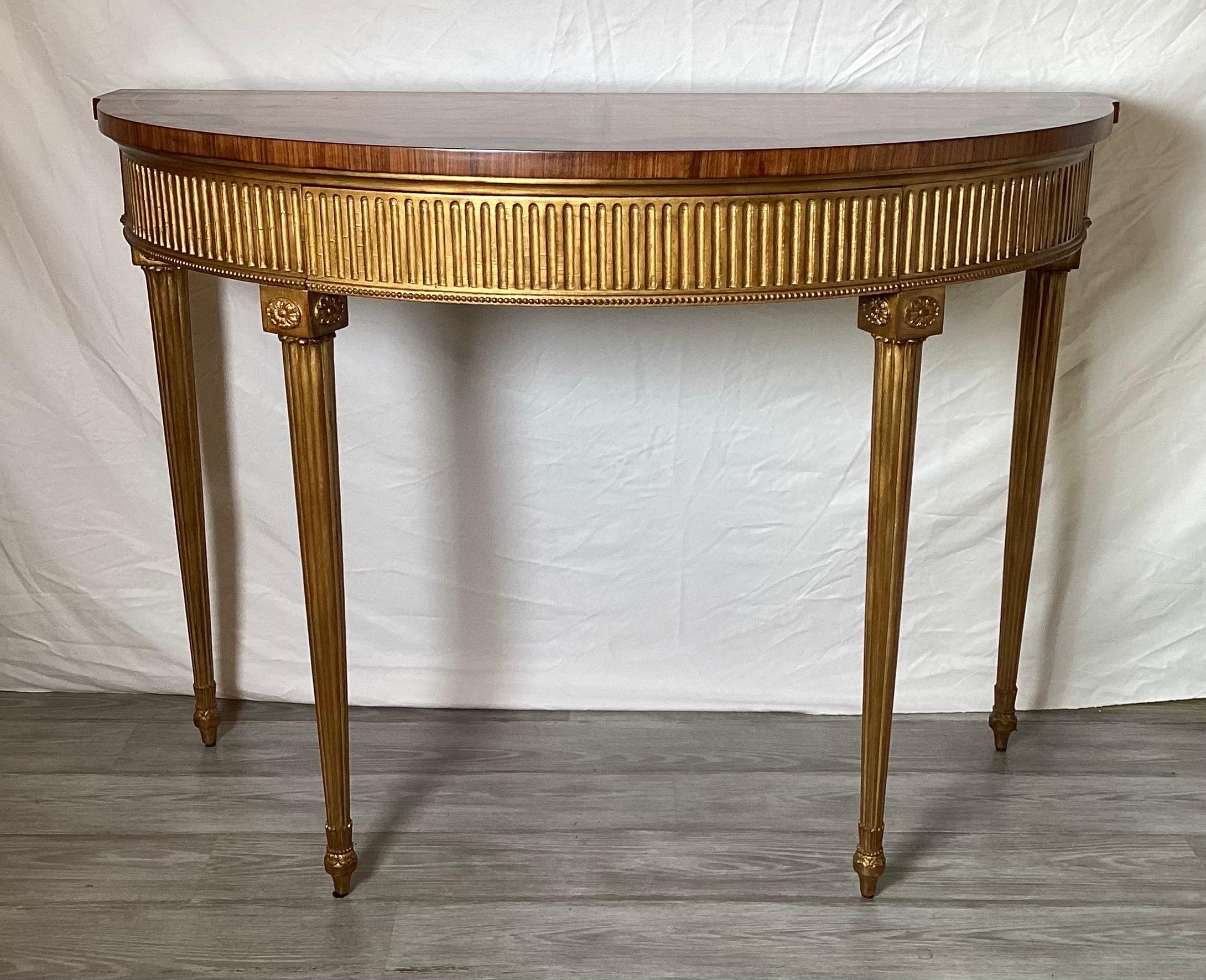 An elegant giltwood and inlaid Demilune console table by Theodore Alexander.  The giltwood apron and legs with fluted detailing with a concealed central drawer.  The top with an Adam style mahogany and satinwood inlaid design.  The generous size is