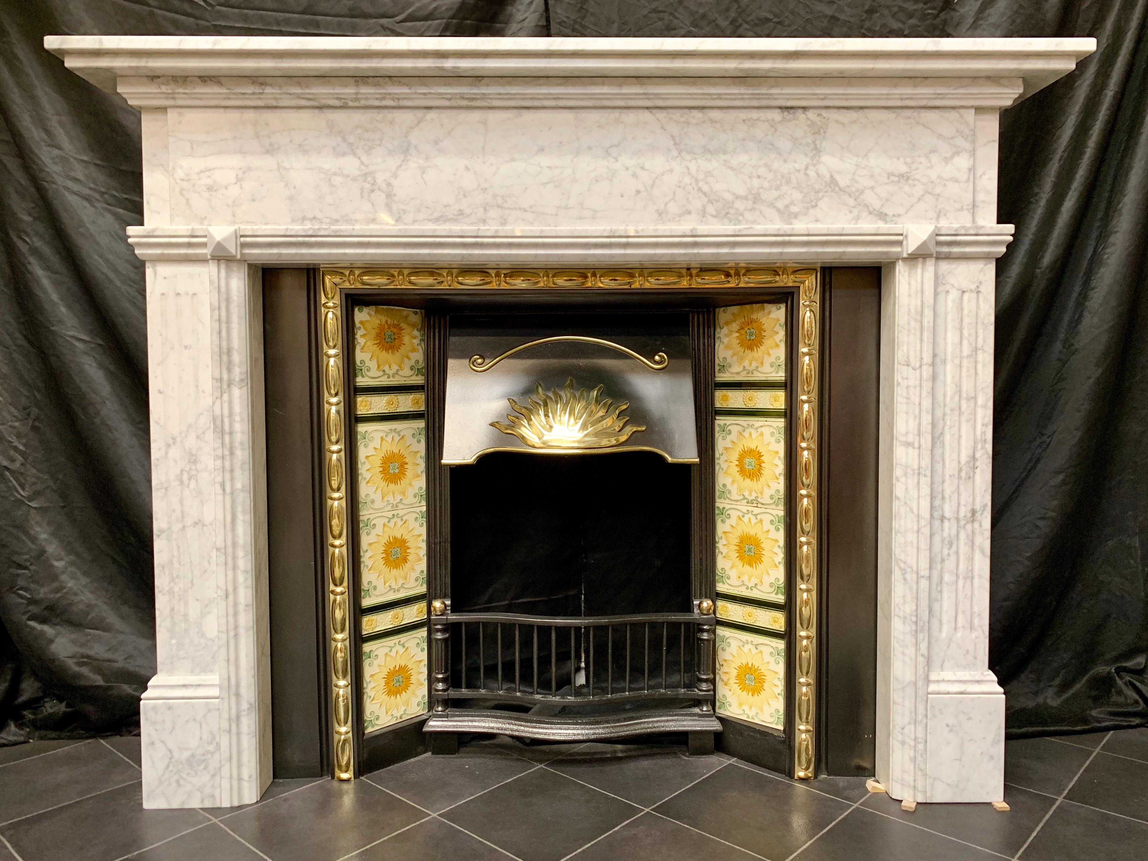 A charming and well proportioned aged Victorian style carved Carrara marble fireplace surround. A double stepped top sits above an unadorned frieze, flanked by tall fluted jambs with a carved opening moulding and splayed blocks to corners, all