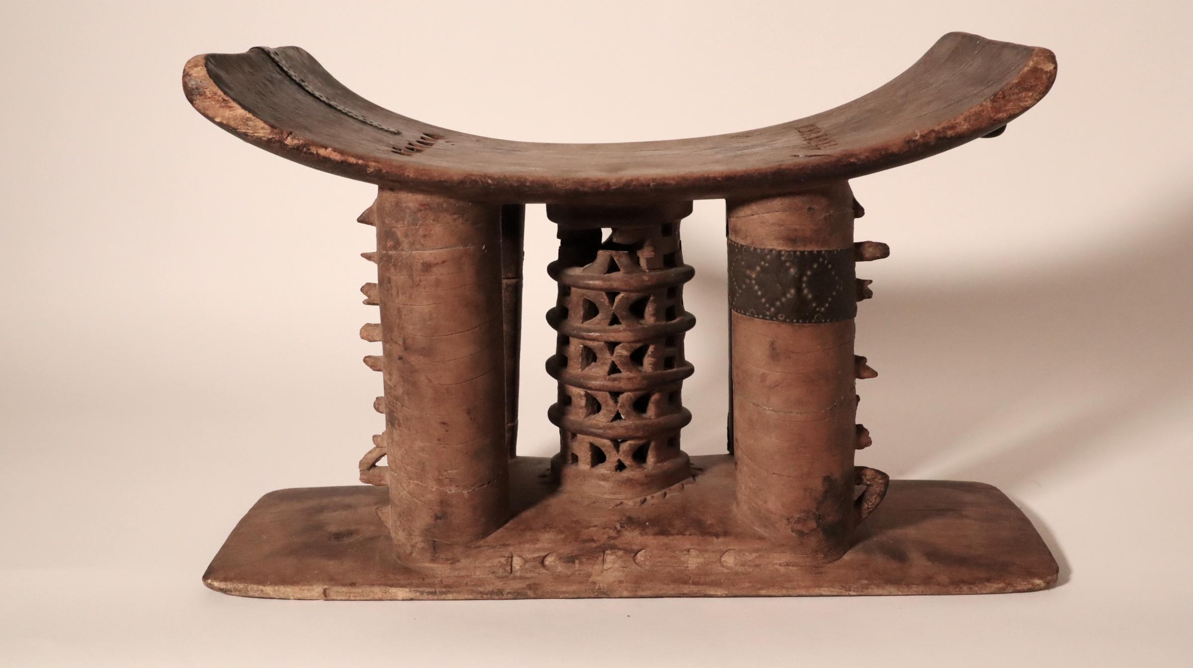 The stool was loved by its owner, repaired in several places with decorated metal straps. It has an openwork, tiered lattice central post and four support posts. It has the older style tiered square cut out and wide cross hollow on the underside