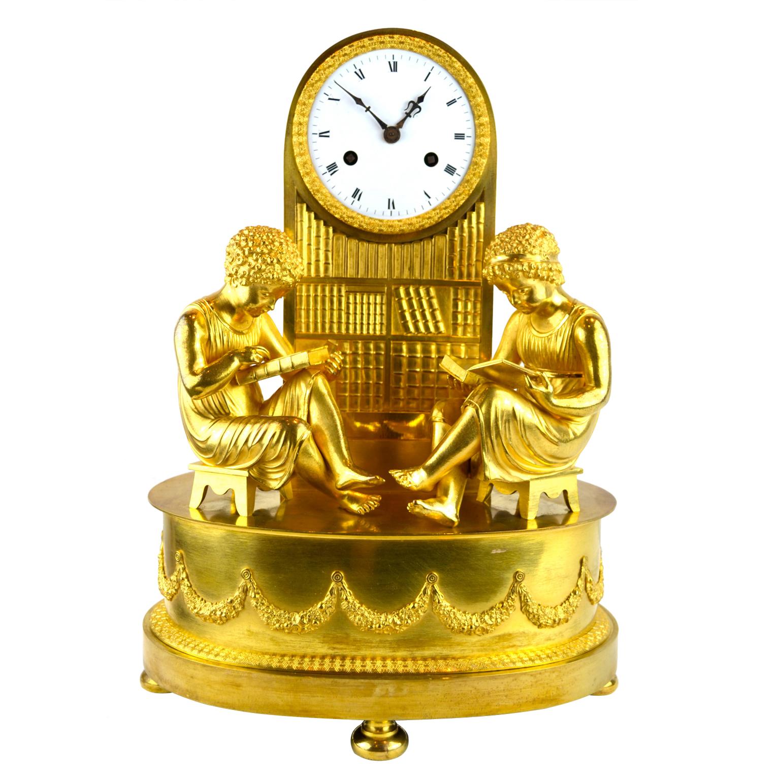 A rare model of a French Empire clock in gilded bronze called The Library, featuring a boy and a girl dressed in classical attire diligently studying in a library, both are sitting on a stool in front of a clock which is integrated into a column