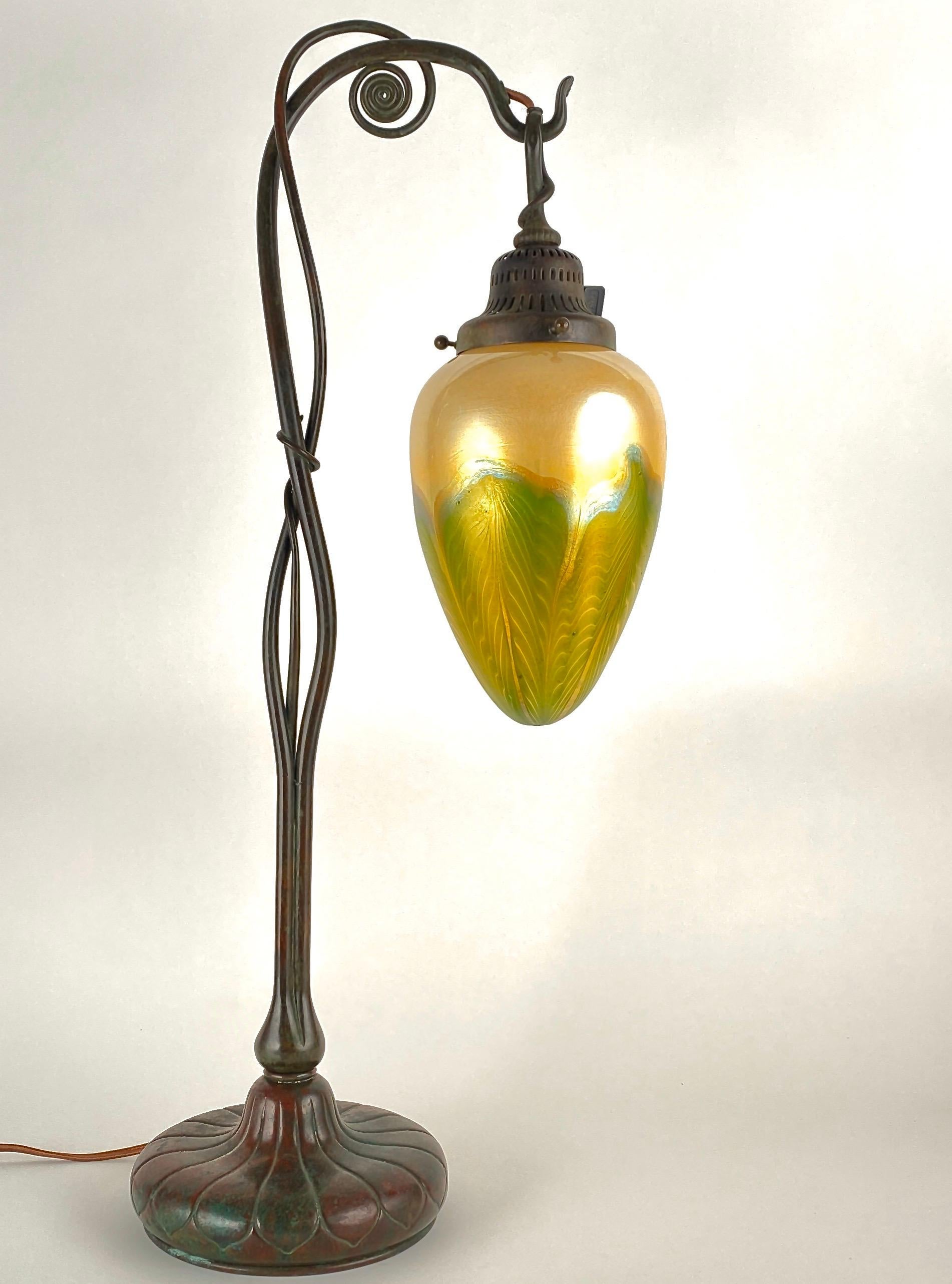 An American Art Nouveau cast bronze and favrile table lamp by, Tiffany Studios decorated with a swivel organically decorated patinated and cast bronze base with a decorated ribbed base. The base is adorned with a blown glass Tiffany Favrile “pulled