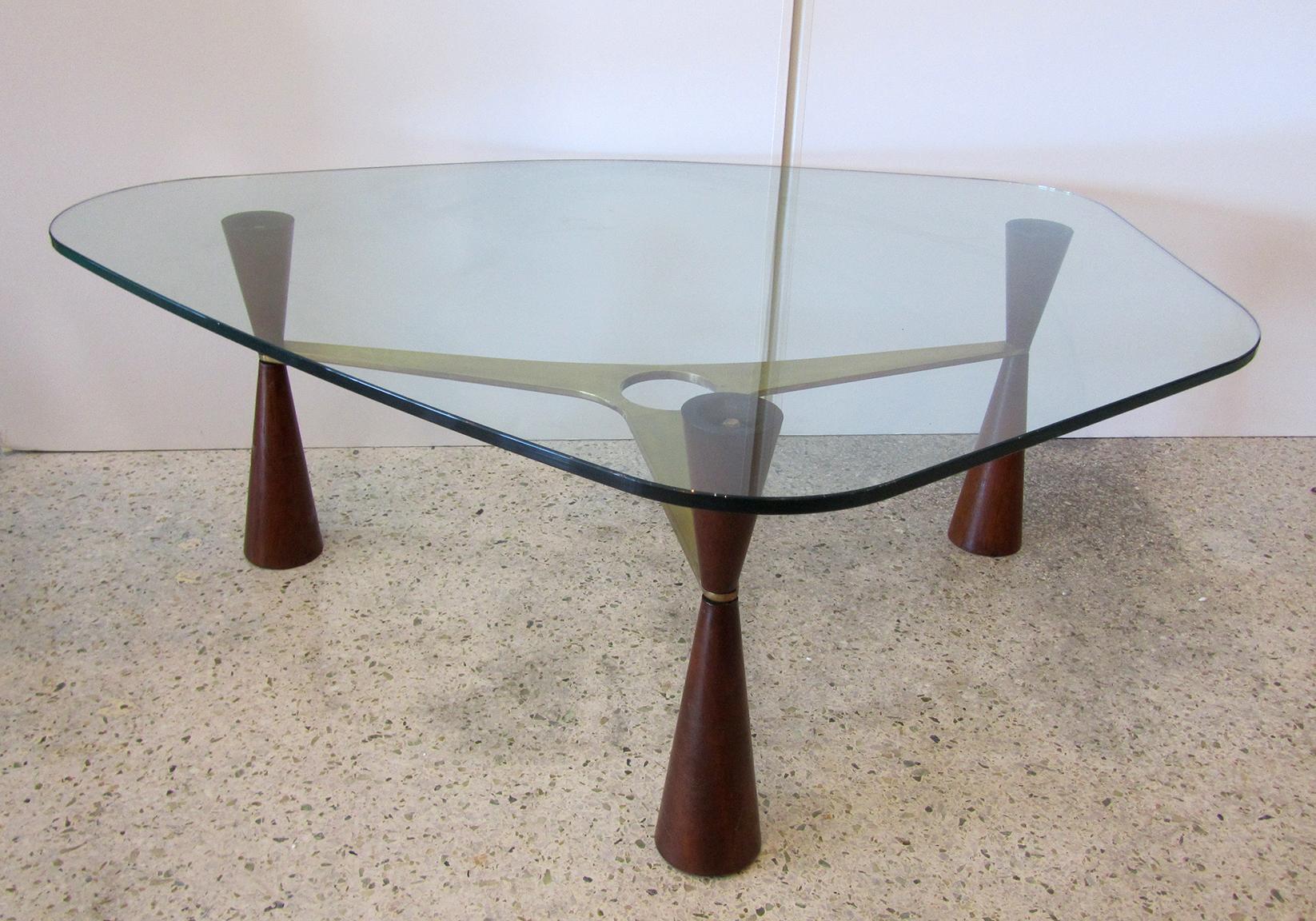 A beautiful and unusual coffee table by Ed Wormley for Dunbar. A freeform glass top sits on top of 3 tapering legs divided by a brass stretcher with a circular cutout. There is beautiful brass hardware throughout the piece. Dimensions of base alone: