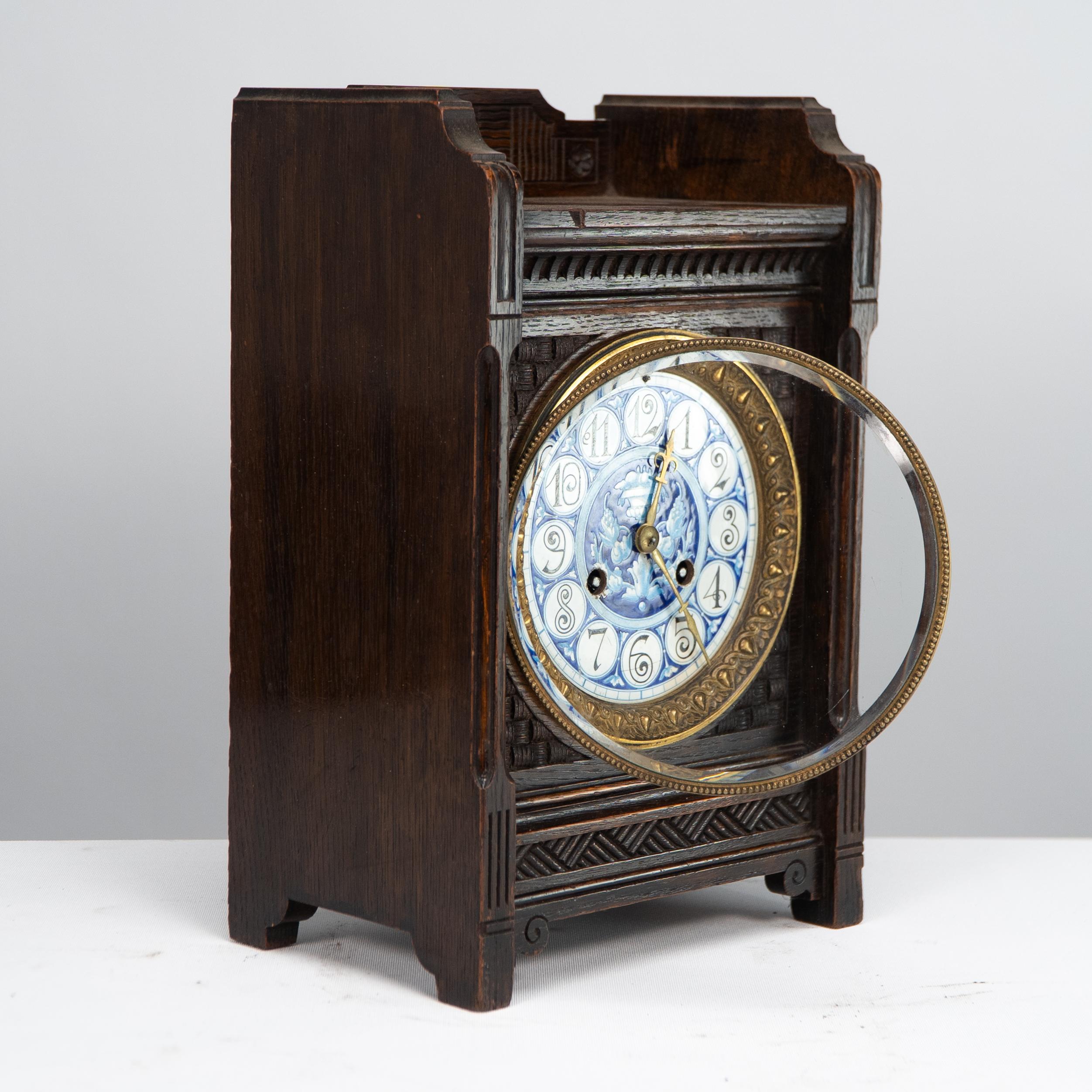 An Anglo-Japanese oak mantel clock with a handpainted blue floral dial surrounded by a floral gilt brass outer ring, the top front and lower section carved with stylized Japanese basket weave decoration, and incised details to the front upright and