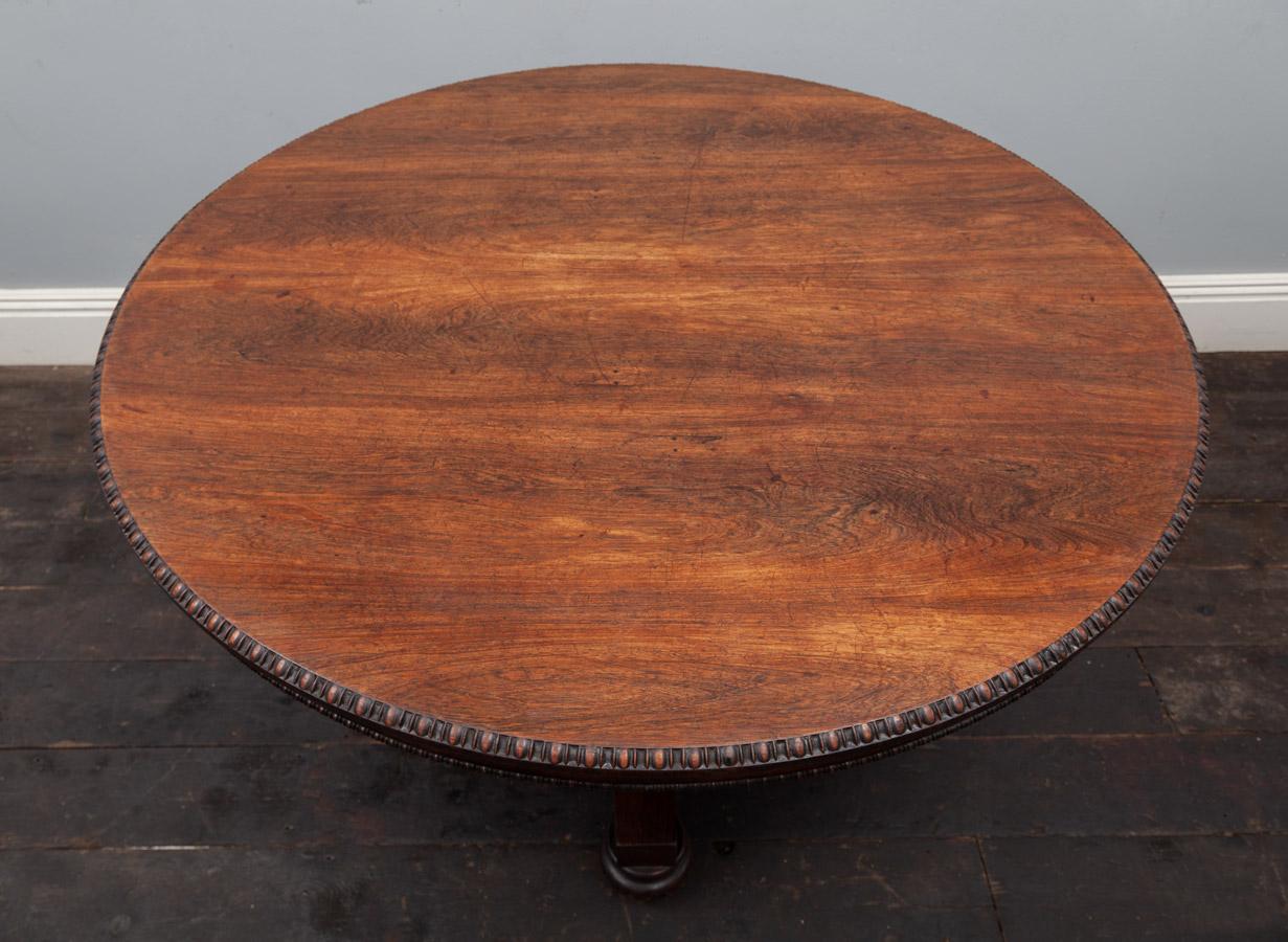 An antique rosewood circular tilt-top table from the George IV period. The solid rosewood top with carved bead and reel rim, below this a plain frieze with a smaller carved bead and real moulding. The curricular centre pillar rests on a triform base