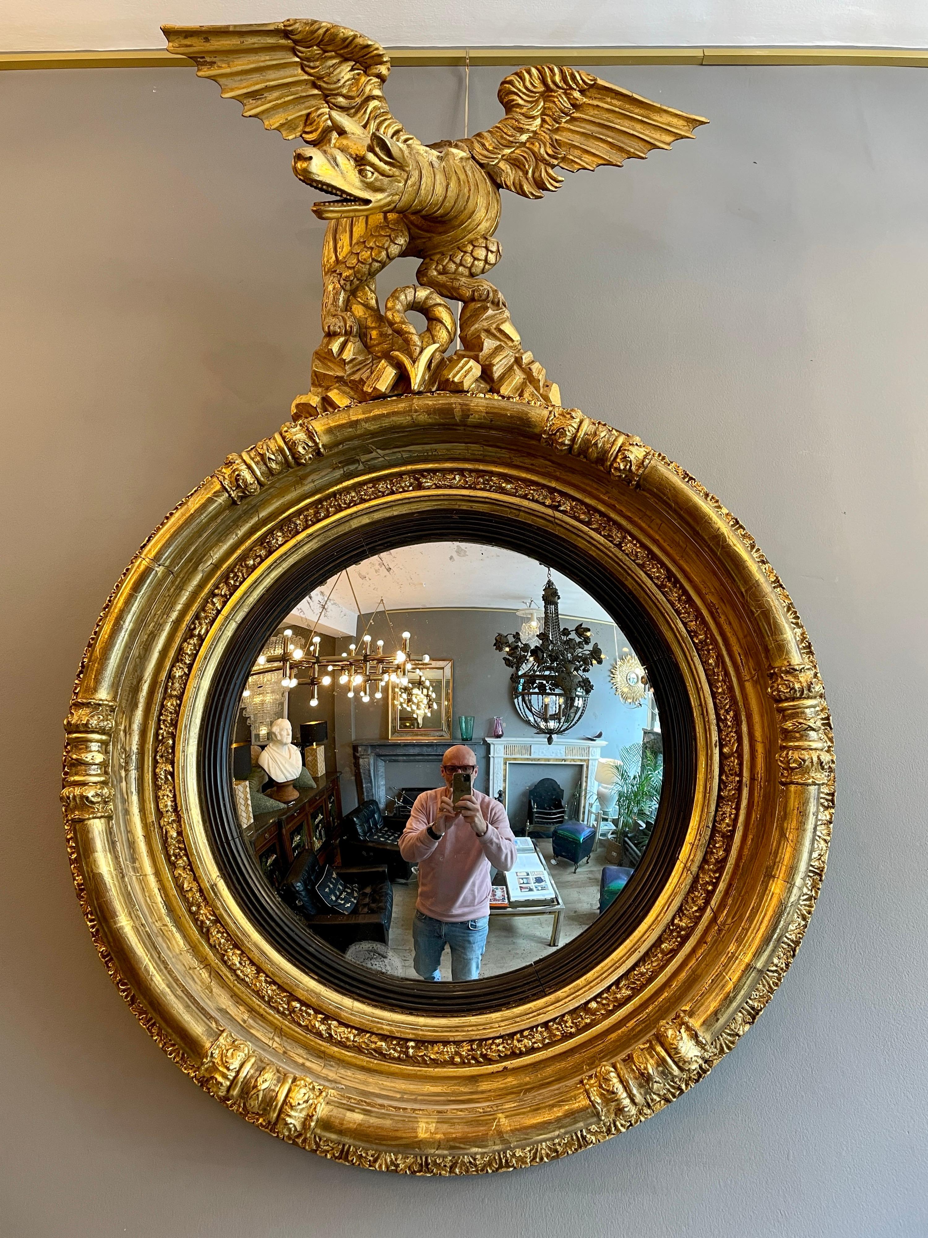 Exceptionnel miroir convexe d'époque Régence en bois sculpté, doré et gesso, vers 1810. Le fronton en bois sculpté représente un dragon perché, aux ailes déployées et à la queue de serpent enroulée, surmontant un cadre profondément moulé et orné,