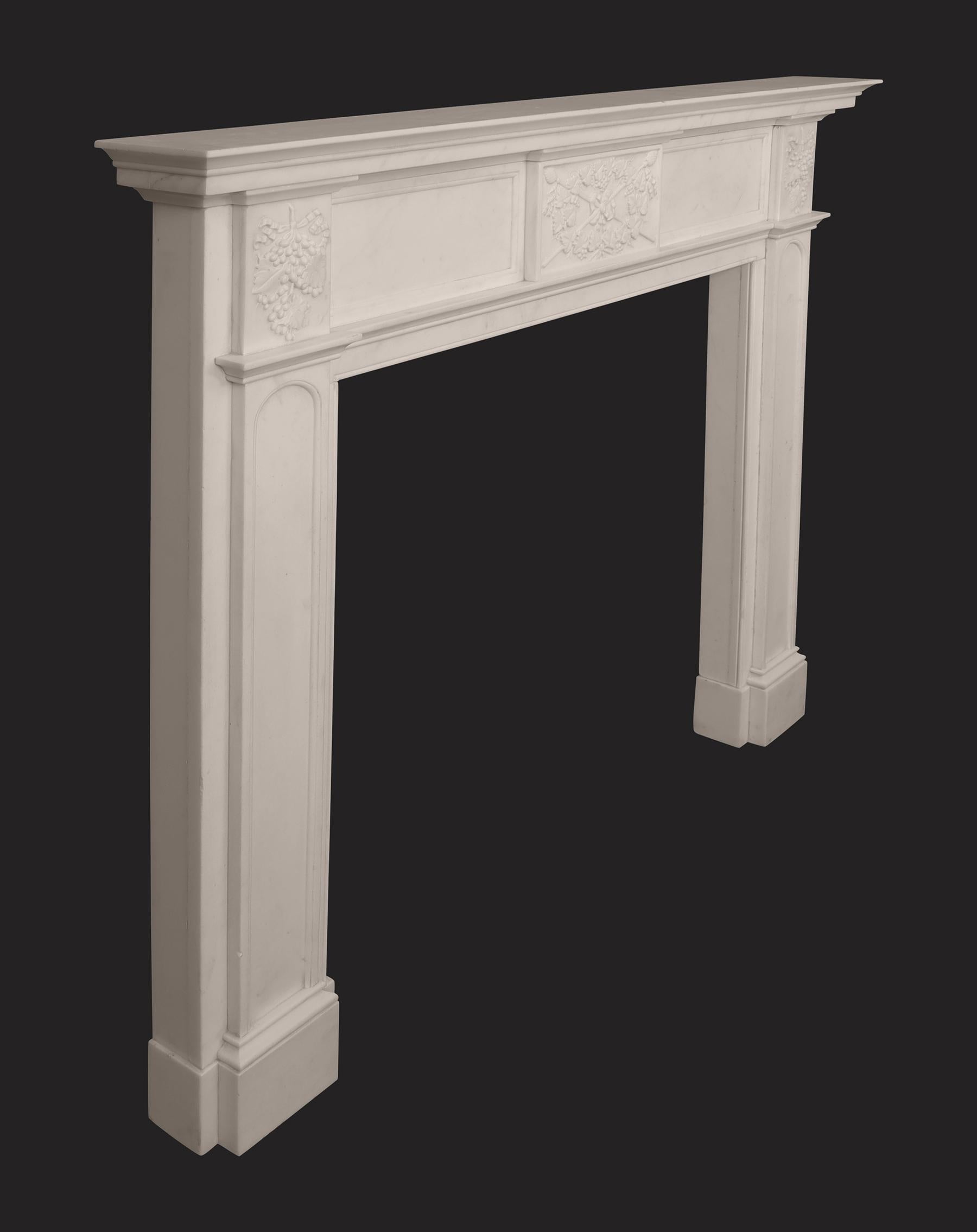An antique English marble fireplace circa 1830-1840 In Italian white marble with panelled frieze and arched panelled jambs on plinths. The central tablet depicts crossed spears, with ribbons and vine wreath under a finely moulded shelf. The corner