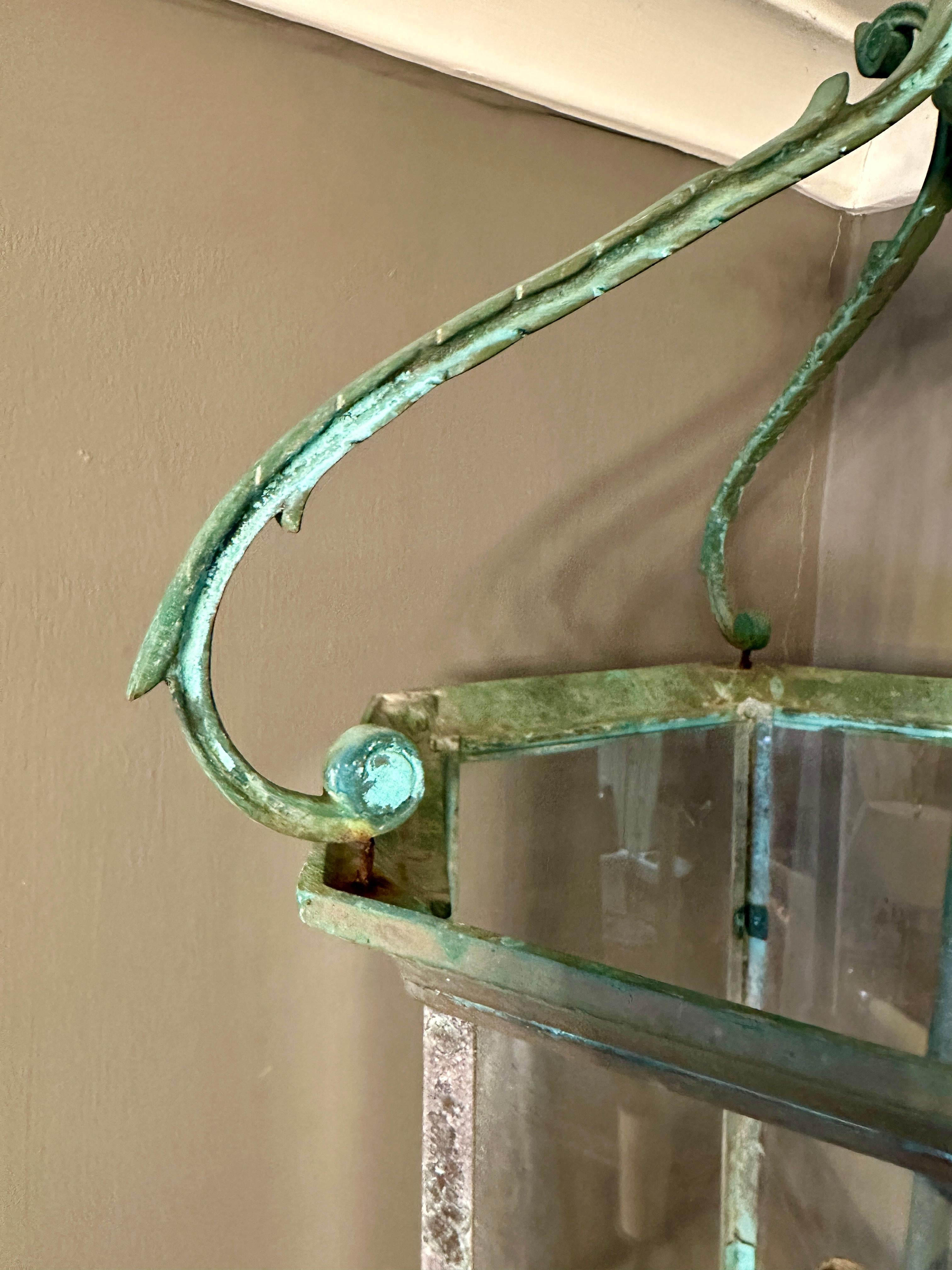 Une grande lanterne en bronze patinée naturellement au vert-de-gris, de forme hexagonale, avec des plaques de verre transparent et une simple branche à trois bras à l'intérieur. L'anneau et la sphère supportent trois bras feuillagés à enroulement