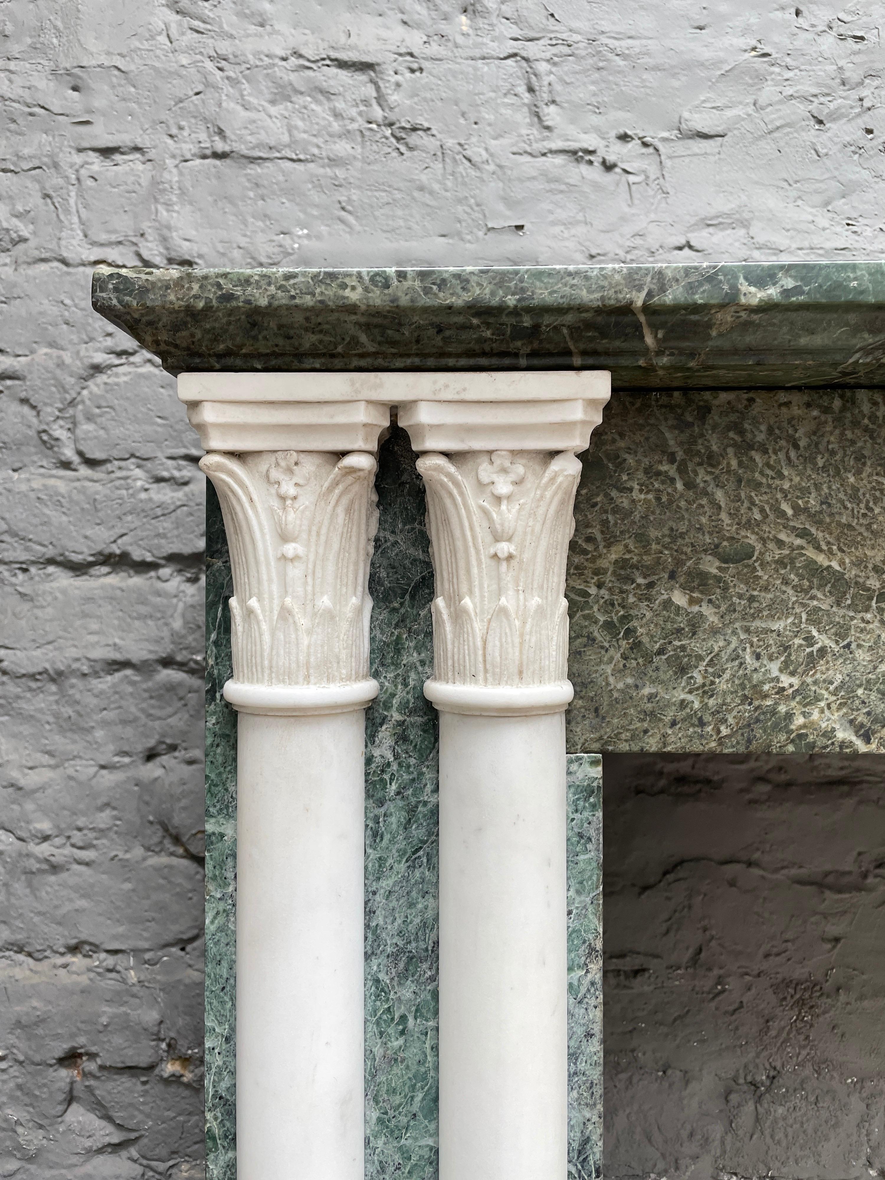 Ein antiker Kamin im Empire-Stil aus Verdi-Antico-Marmor mit halb freistehenden Säulen aus statuarischem weißem Marmor an den Jambs. Die Kapitelle in korinthischer Manier sind ebenfalls aus statuarischem Marmor gefertigt. Ein einfacher Fries und ein