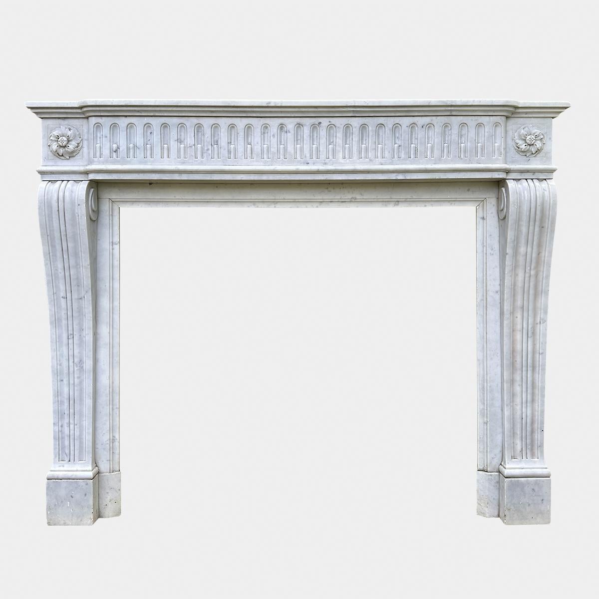 An antique French Louis XVI style fireplace in Italian Carrara marble. The fluted console jambs with carved square patarae corner blocks and bowed running frieze of stop fluted design. The shelf again bow fronted conforming with the frieze. Circa