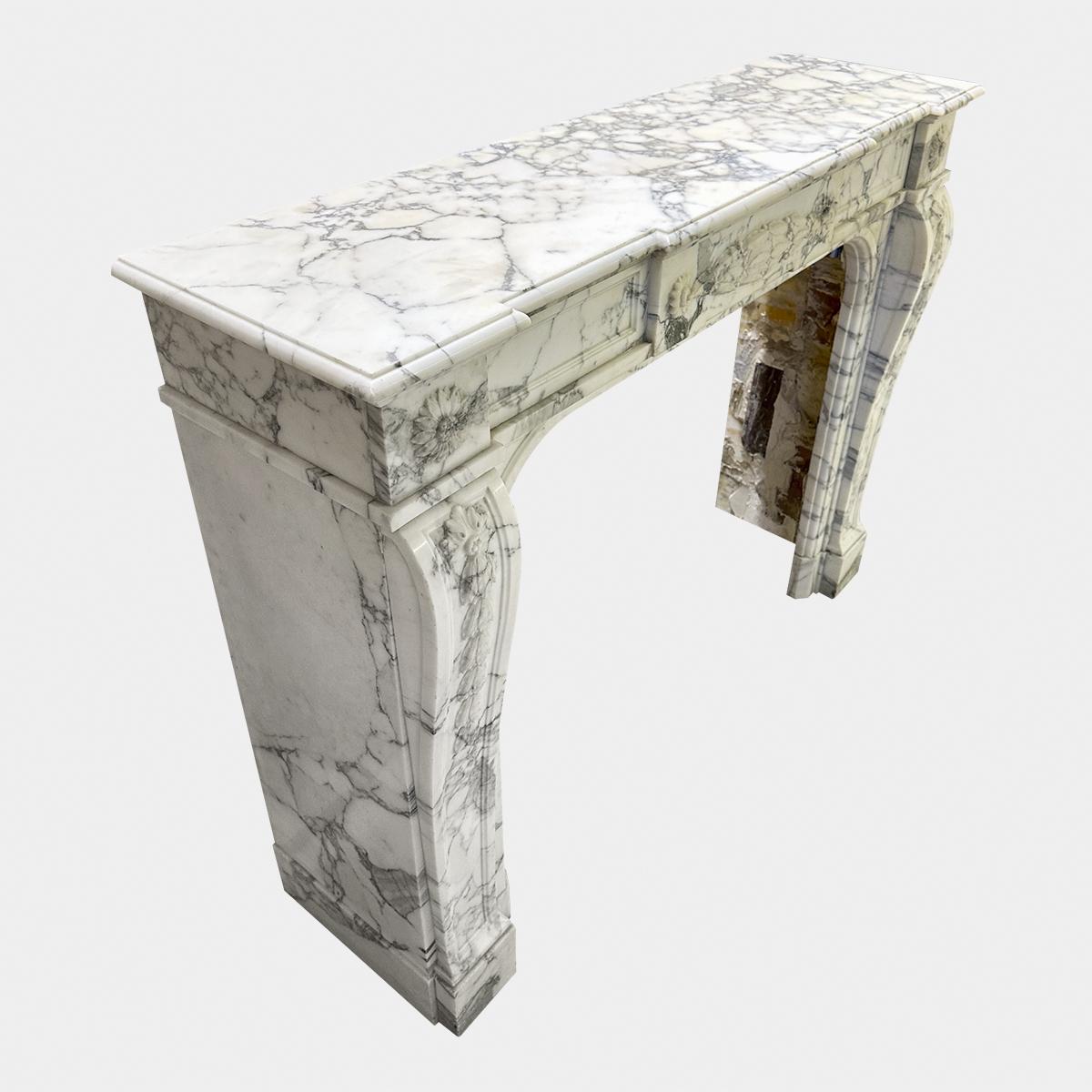 Cheminée ancienne de style Louis XVI en marbre italien Arabescato, datant du milieu du XIXe siècle. Les montants de la console sont sculptés de lauriers et de baies descendant d'une rosace circulaire sculptée de fleurs. La frise est ornée d'une