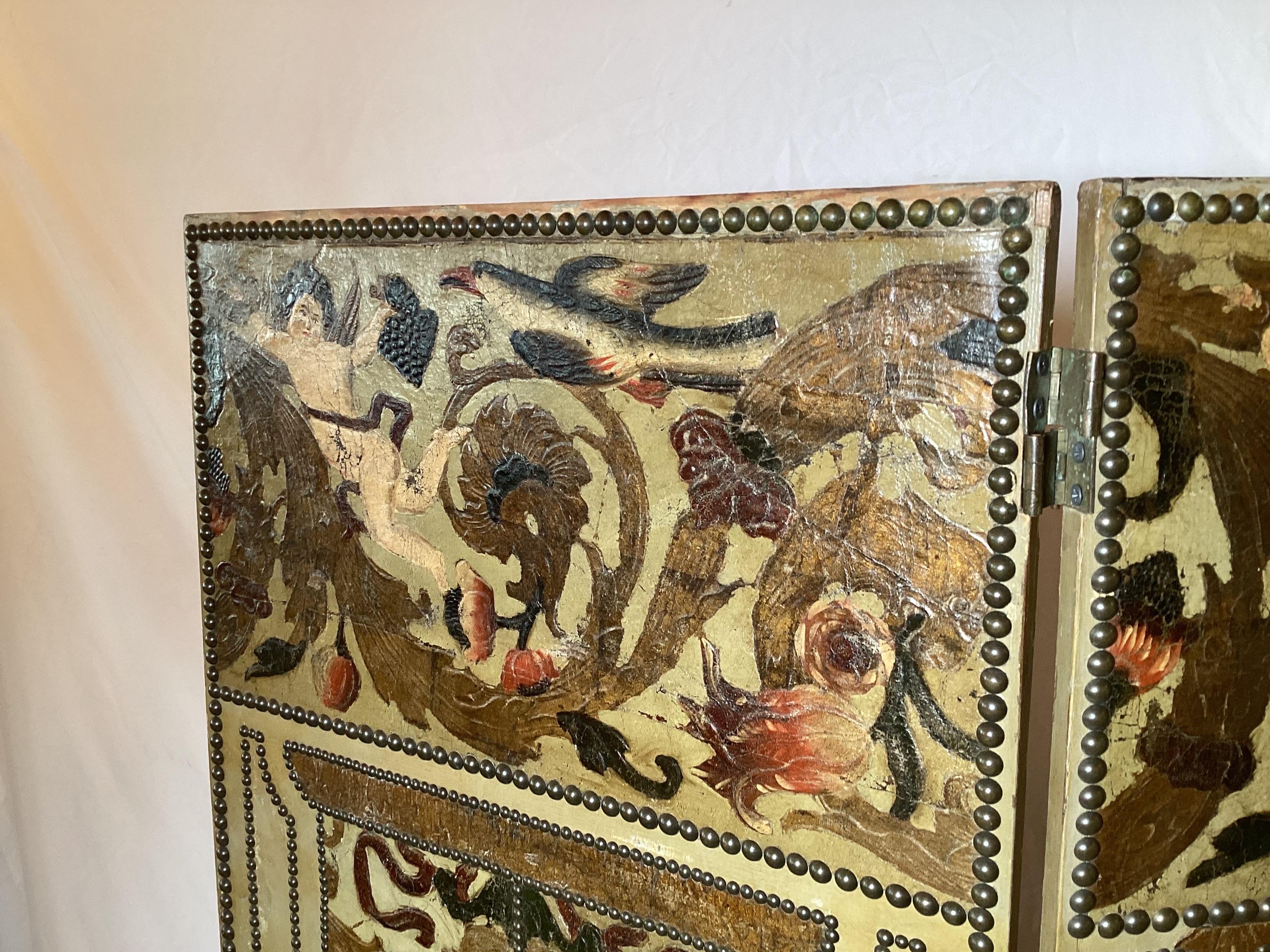 A three panel hand painted and hand tooled Italian leather screen. Decorated with cupids, birds, and floral design with nail head trim details . The vibrantly colored front the back with a trellis pattern with brass nail head trim. folds to 20