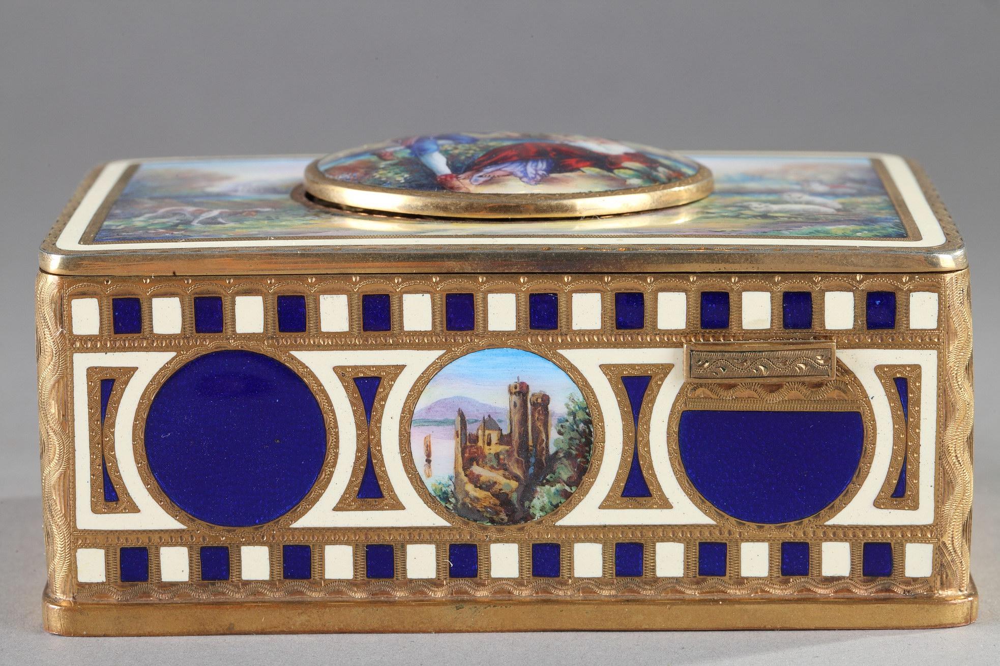 Rectangular music box in gilded brass and polychrome enamel. The music box is adorned on the sides with translucent blue enamel panels and medallions depicting representing an alpine lakescape with medieval castles. The lid is decorated with a scene