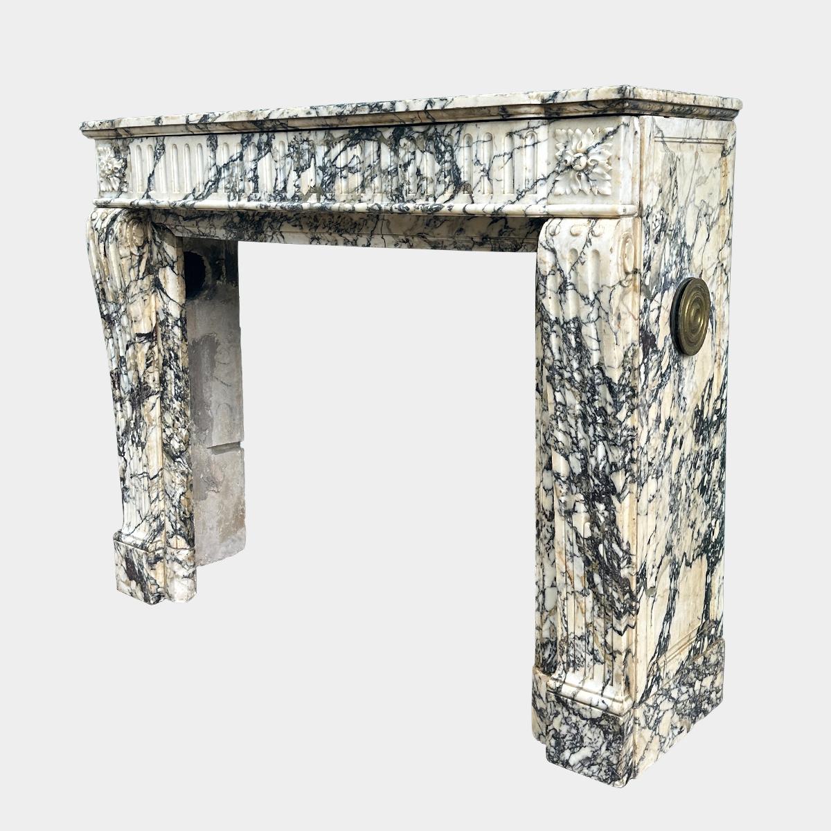 Un camino ben proporzionato in stile Luigi XVI, scolpito in marmo di Serravezza molto variegato. Gli stipiti della console sono scanalati con ornamenti a semi-sfera. Il fregio frontale ad arco è scanalato con blocchi terminali di Patarae quadrate