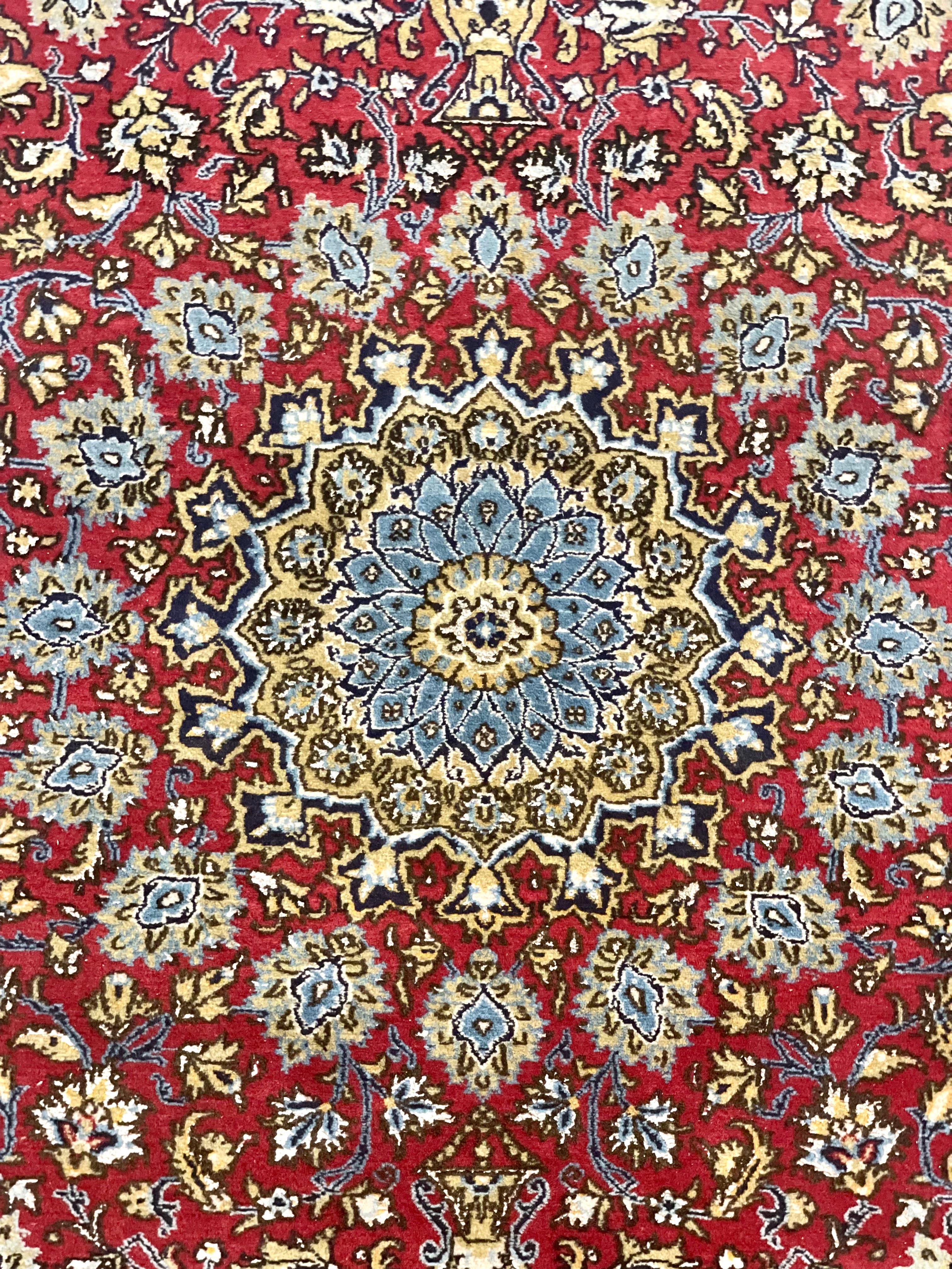 A Persian 'Qum' or 'Ghoum' rug hand-woven in wool and silk, featuring a central star- shaped medallion in vivid hues of red and blue, from which rosettes and flowers radiate outwards towards a sandy-coloured border. Prized for the complexity and