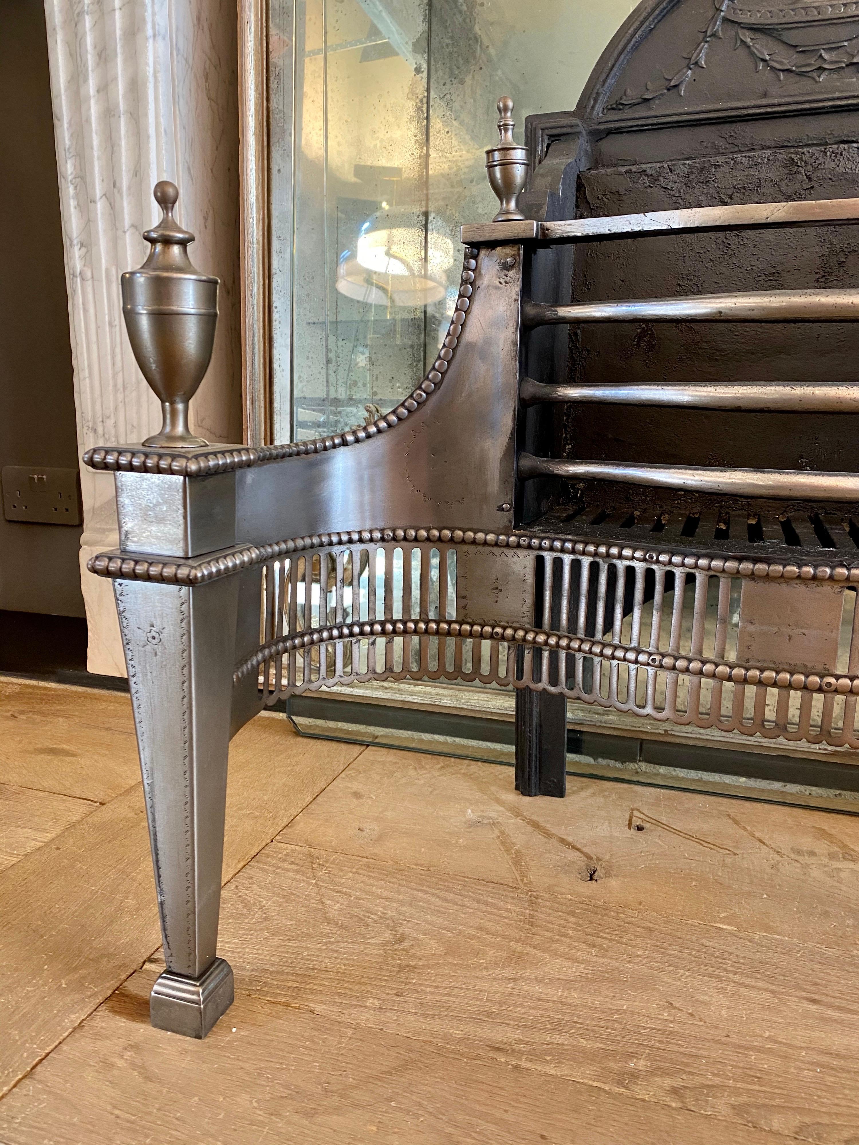 A large polished steel fire grate by Thomas Elsley London. The serpentine fluted pierced fretwork having beaded edging, flanked by engraved tapering supports with classical urn finials. The beaded wings and bowed burning bars again with conforming