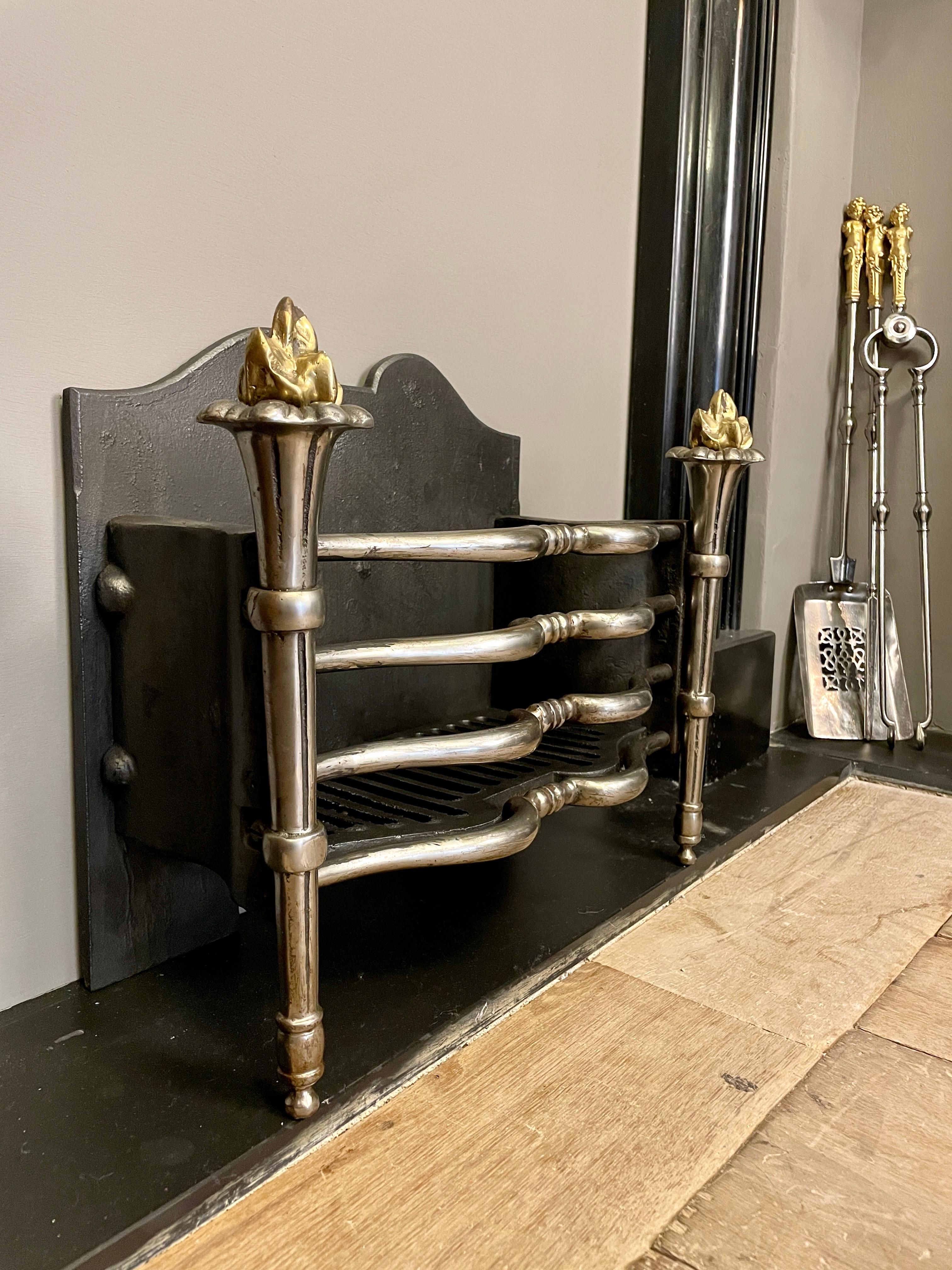 A turn of the 20th century circa 1900 English Brass, steel and cast iron fire grate/basket. The tapering front  torchiere supports with brass flame finials, flanking Archers bow shaped front bars, with conforming shaped cast iron fire back. A good