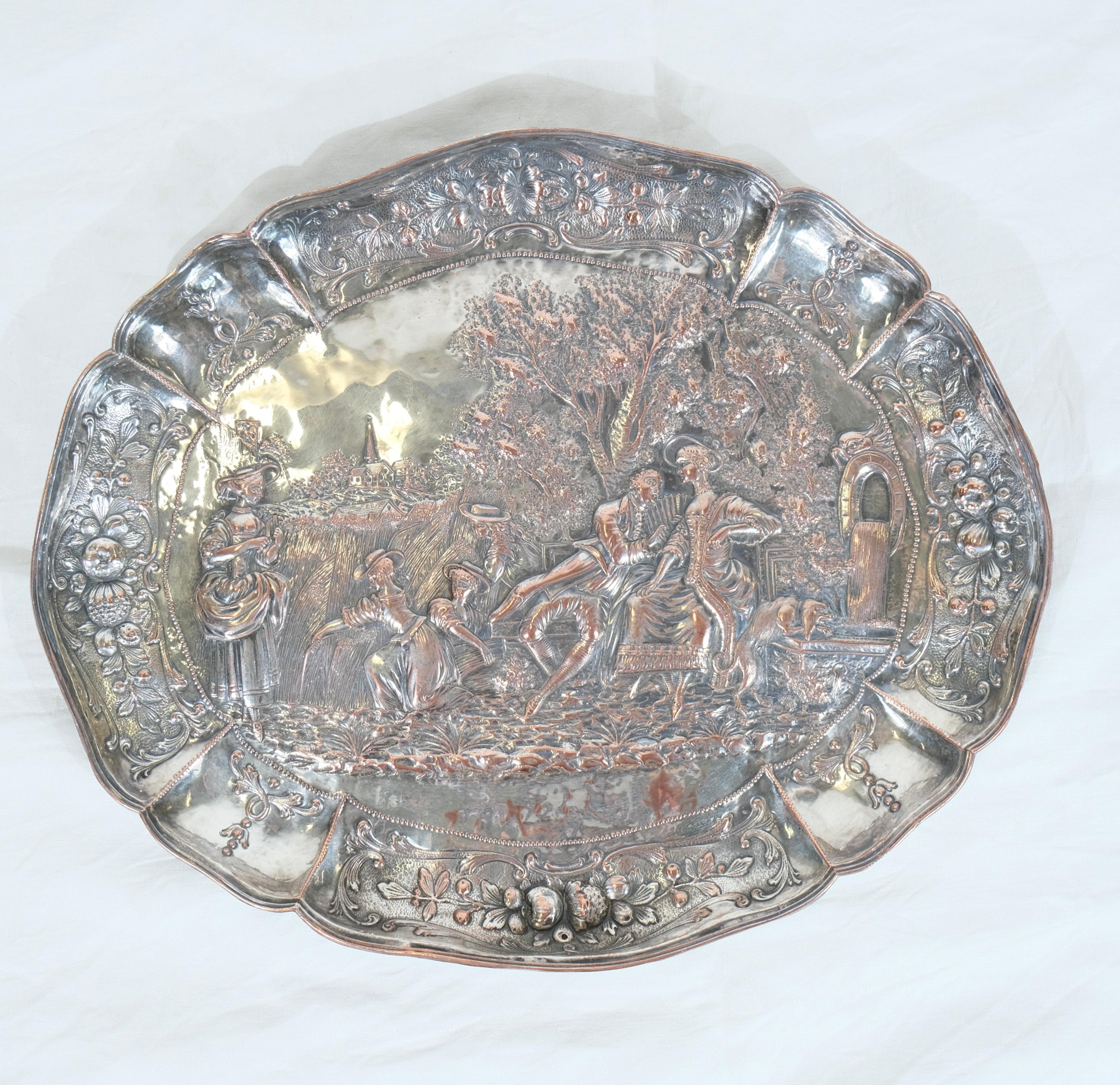 Une grande assiette très décorative en cuivre argenté. La plaque est ornée d'un motif ciselé dans le cuivre. Elle représente une scène romantique avec un homme faisant la cour à une femme près d'une fontaine où un chien s'abreuve. À côté, on
