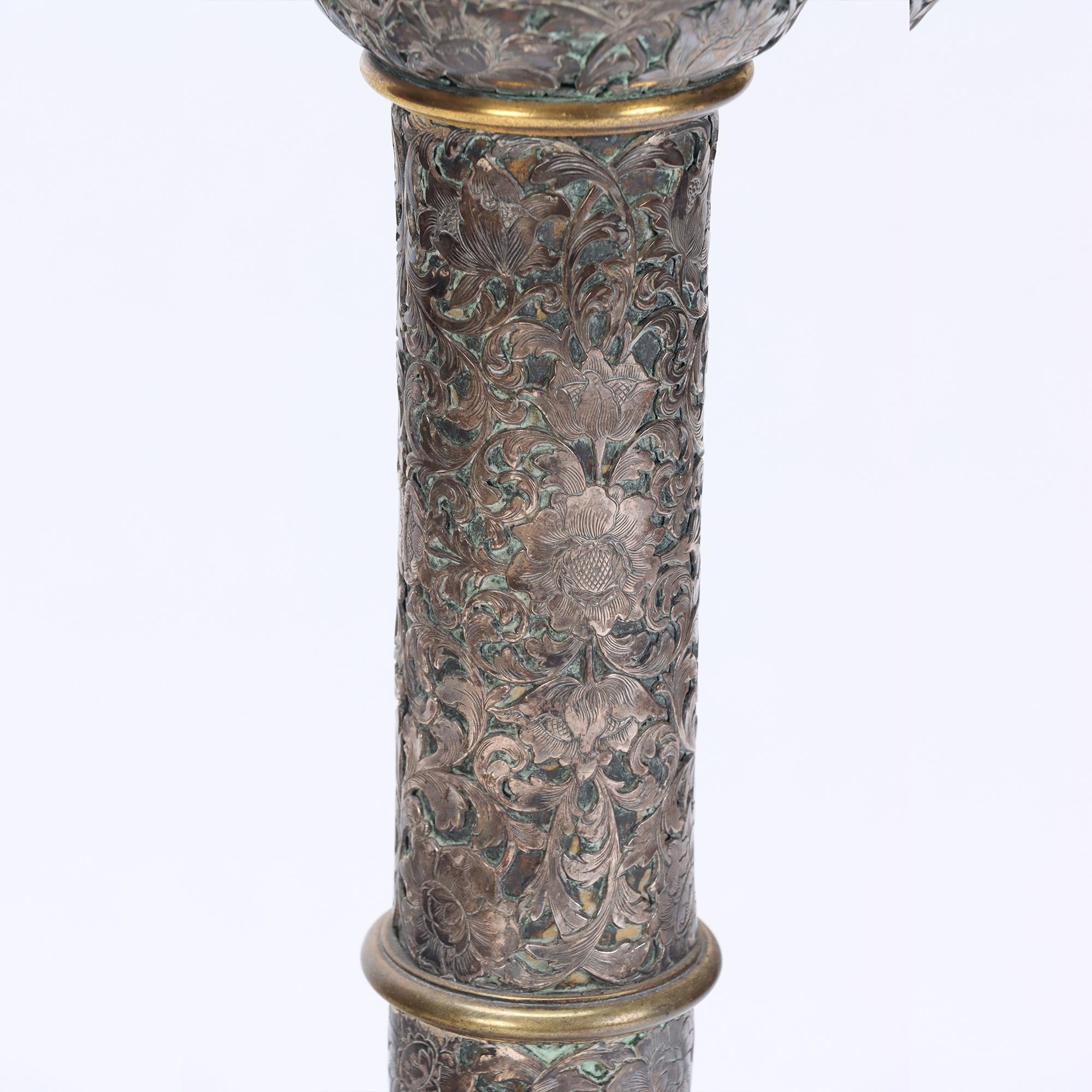 The columnar standard is composed of a series of turned and banded sections, richly chased and engraved with scrolling foliage and floral motifs in high relief. The lamp retains a beautiful aged silvered finish with gilt bronze accents, producing a