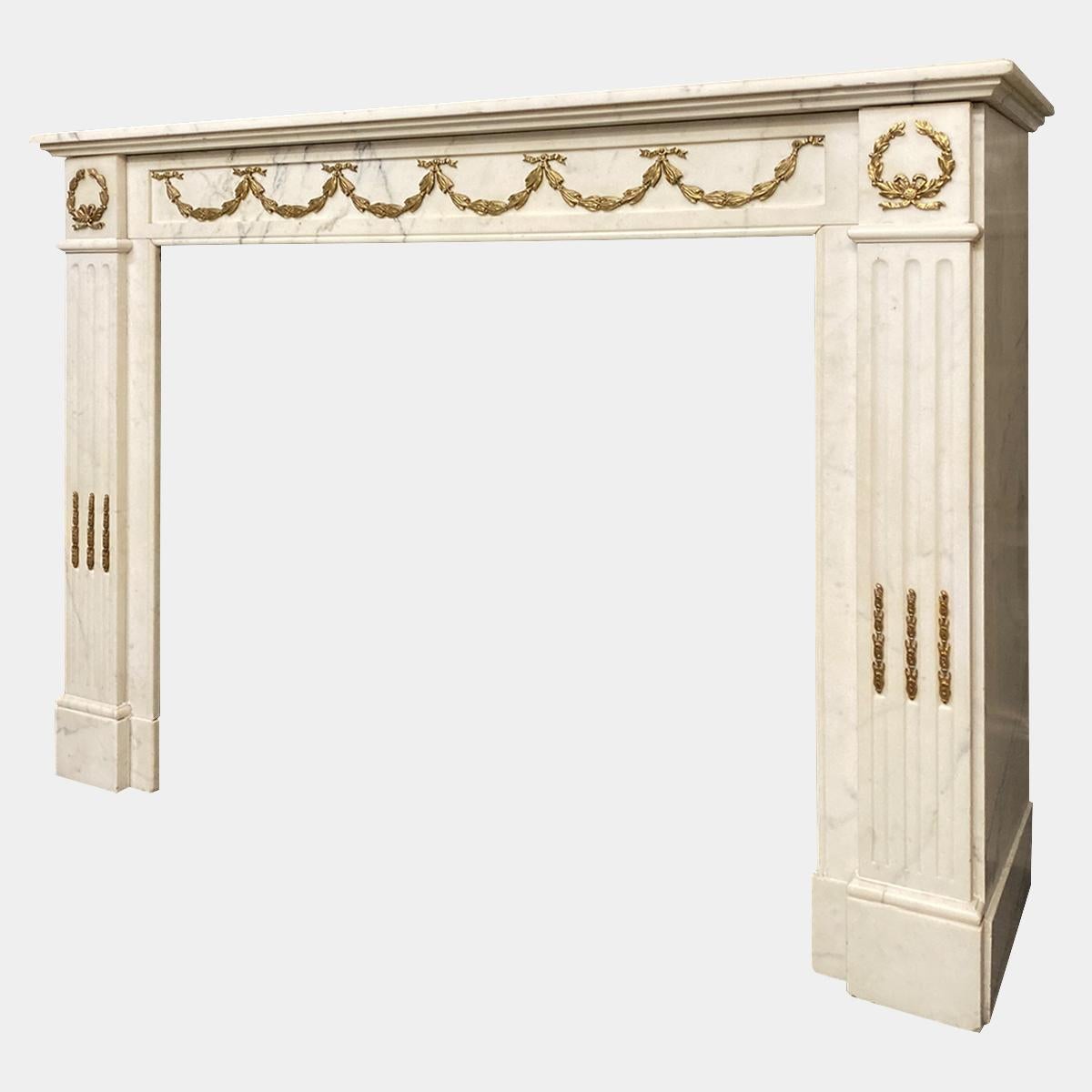 A Statuary white marble and gilt ormolu antique fireplace in the Louis XVI style. The panelled frieze with ormolu bell flower swags, flanked by end blocks of ribbon tied ormolu laurel leafs. The flute jambs with ormolu husks. All beneath a stepped
