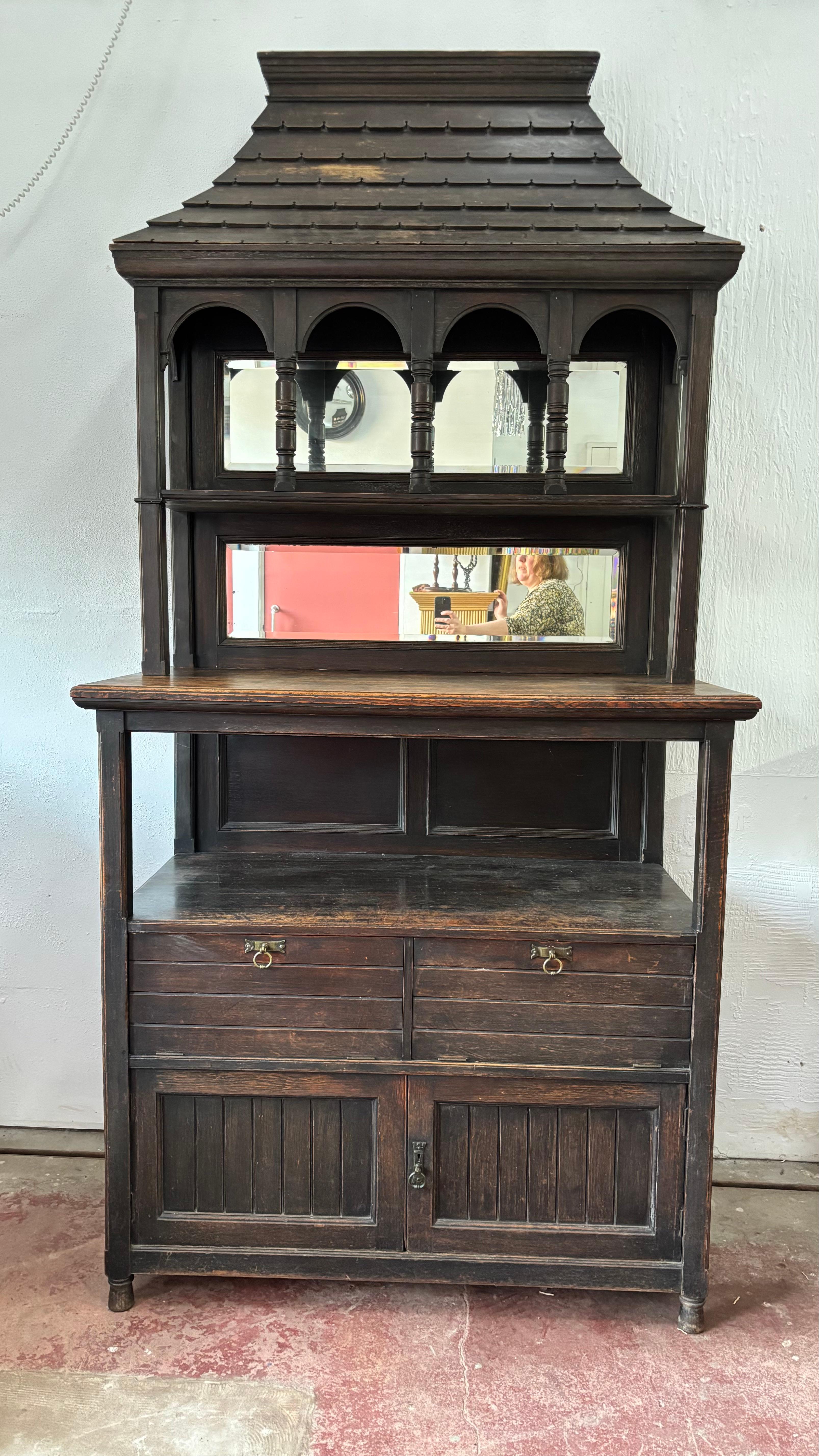 A Unique and special large Arts and Crafts Cabinet. It features an unusual simulated tile pagoda roof, then a top section with mirror back and arched top cabinetry. The middle section is a large shelf, below is an open storage space. Below we have