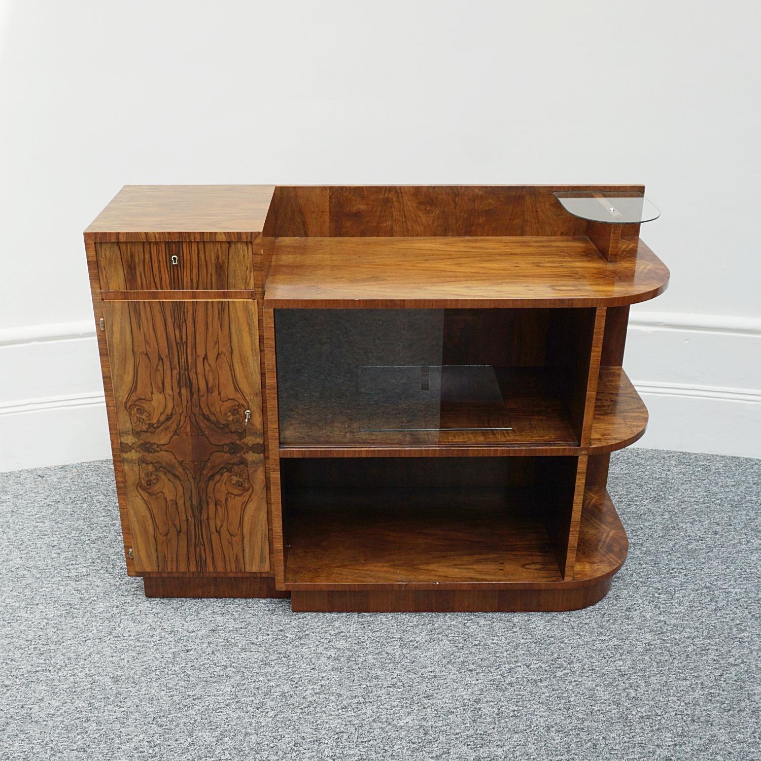 An Art Deco modernist cabinet. Burr walnut with glass sliding front facing doors and three tiered shelving to side. Raised top glass shelf , pull out draw and cupboard with original key. 

Dimensions: H 91.5cm W 120cm D 42cm

Origin: