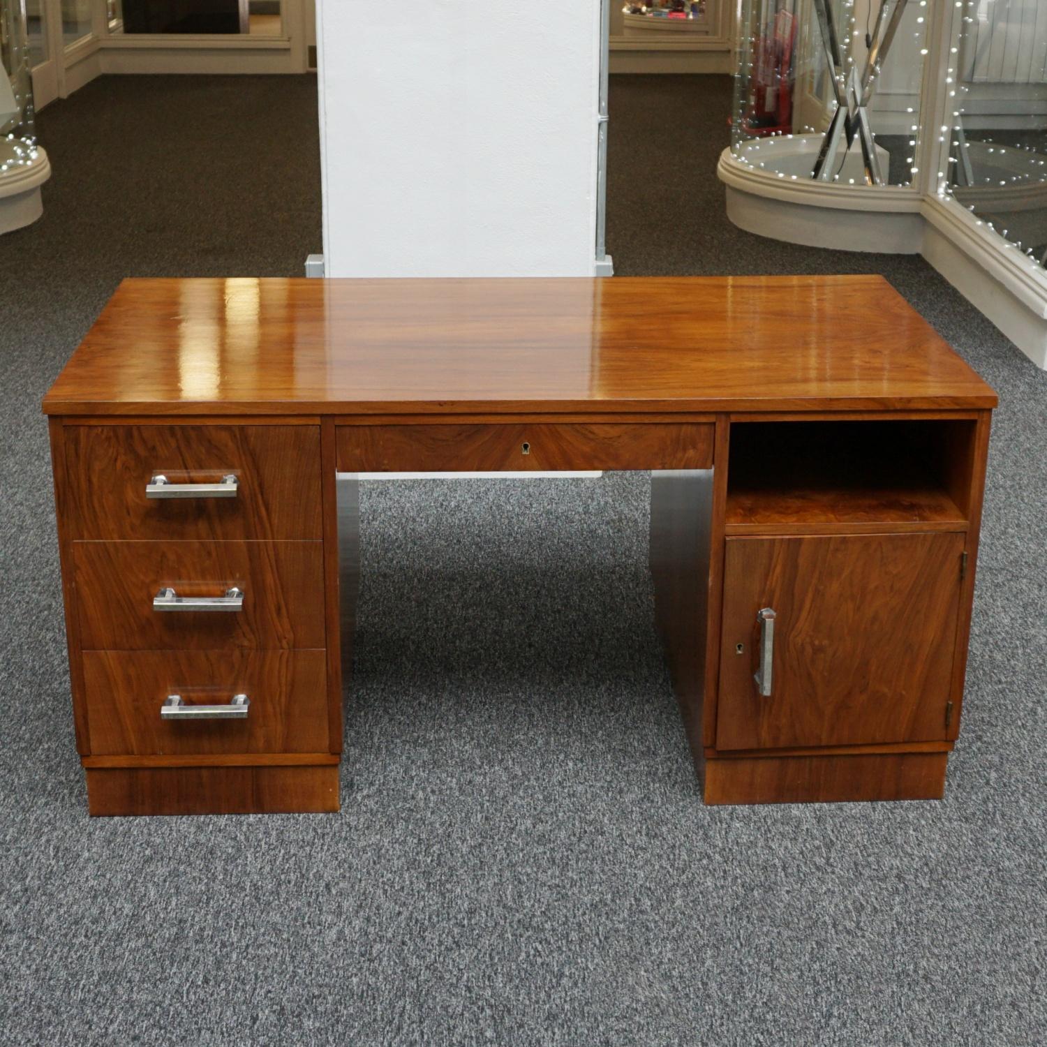An Art Deco Desk. Burr and Figured walnut with original chrome handles. Three deep drawers to the left with a central upper drawer and left hand side open shelf with a lower cupboard opening to reveal inset shelves. Veneered to sides and back.