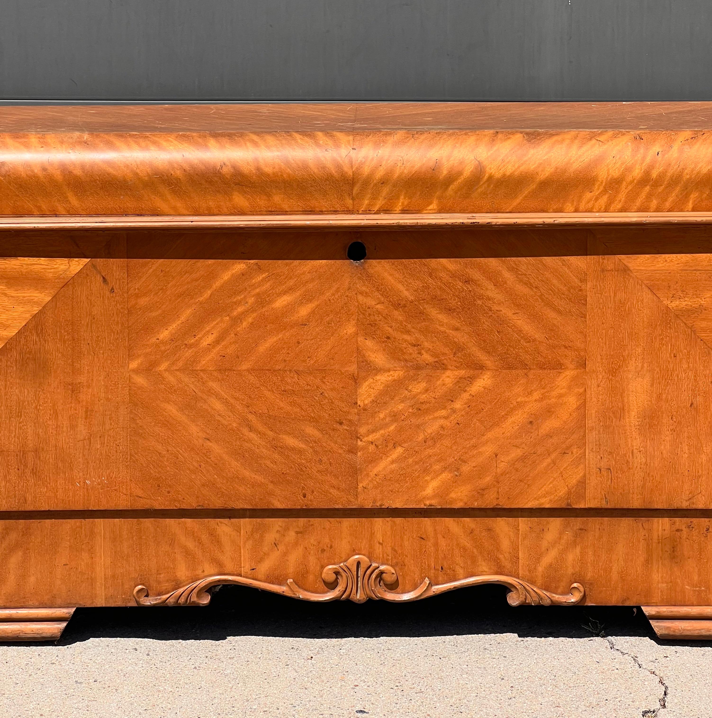 A gorgeous Art Deco waterfall chest by Lane Furniture, circa 1950. Sumptuous shades of wheat and honey interlock in the wood grain to create an almost burl-like effect. The edges have been highly-carved by hand with motifs vaguely reminiscent of Art