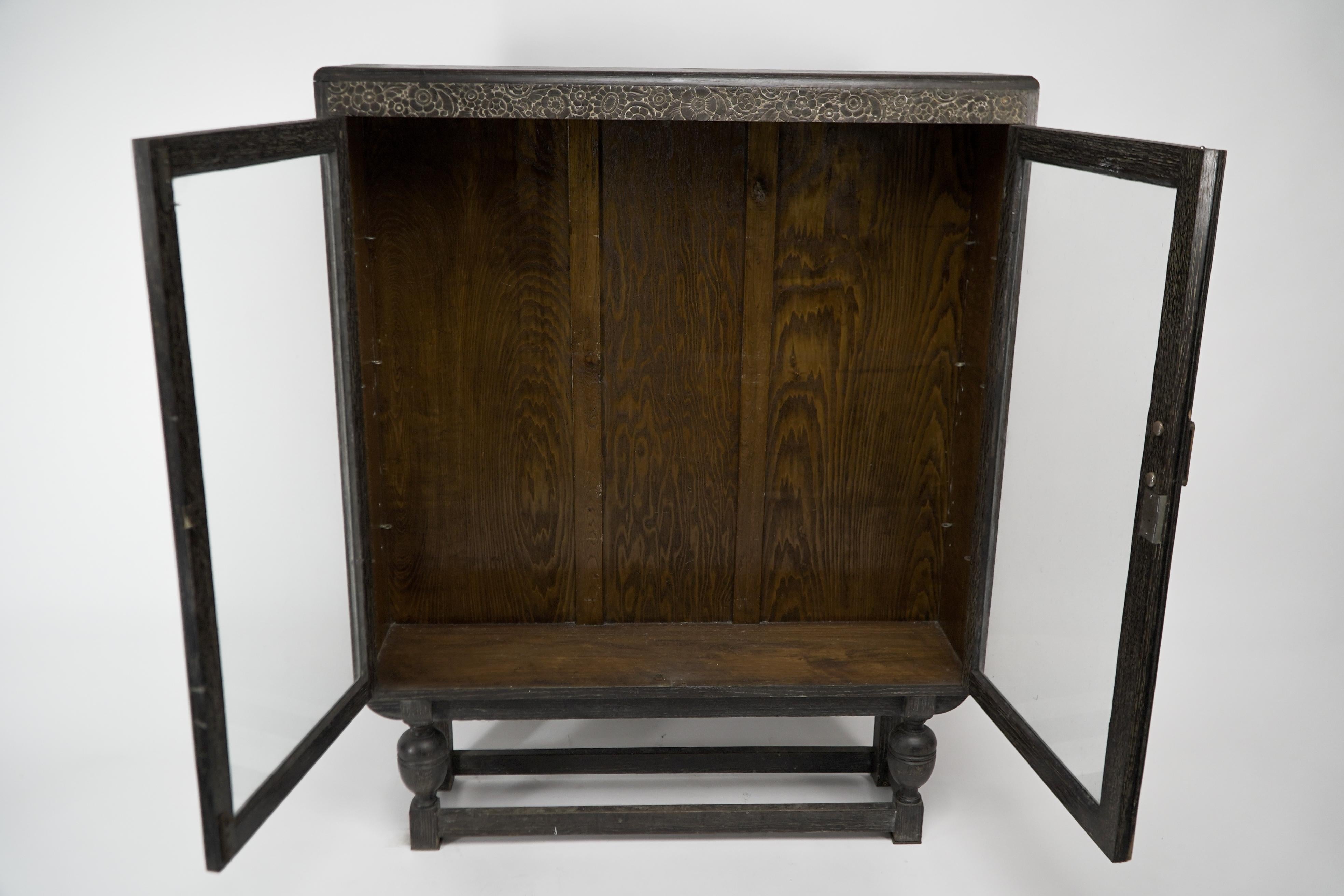 An Art Deco dark oak glazed bookcase with a subtle lime wash to the grain, carved floral decoration to the top and soft rounded edges all over, stood on exaggerated bulbous legs united by straight stretchers and three adjustable shelves (not shown).
