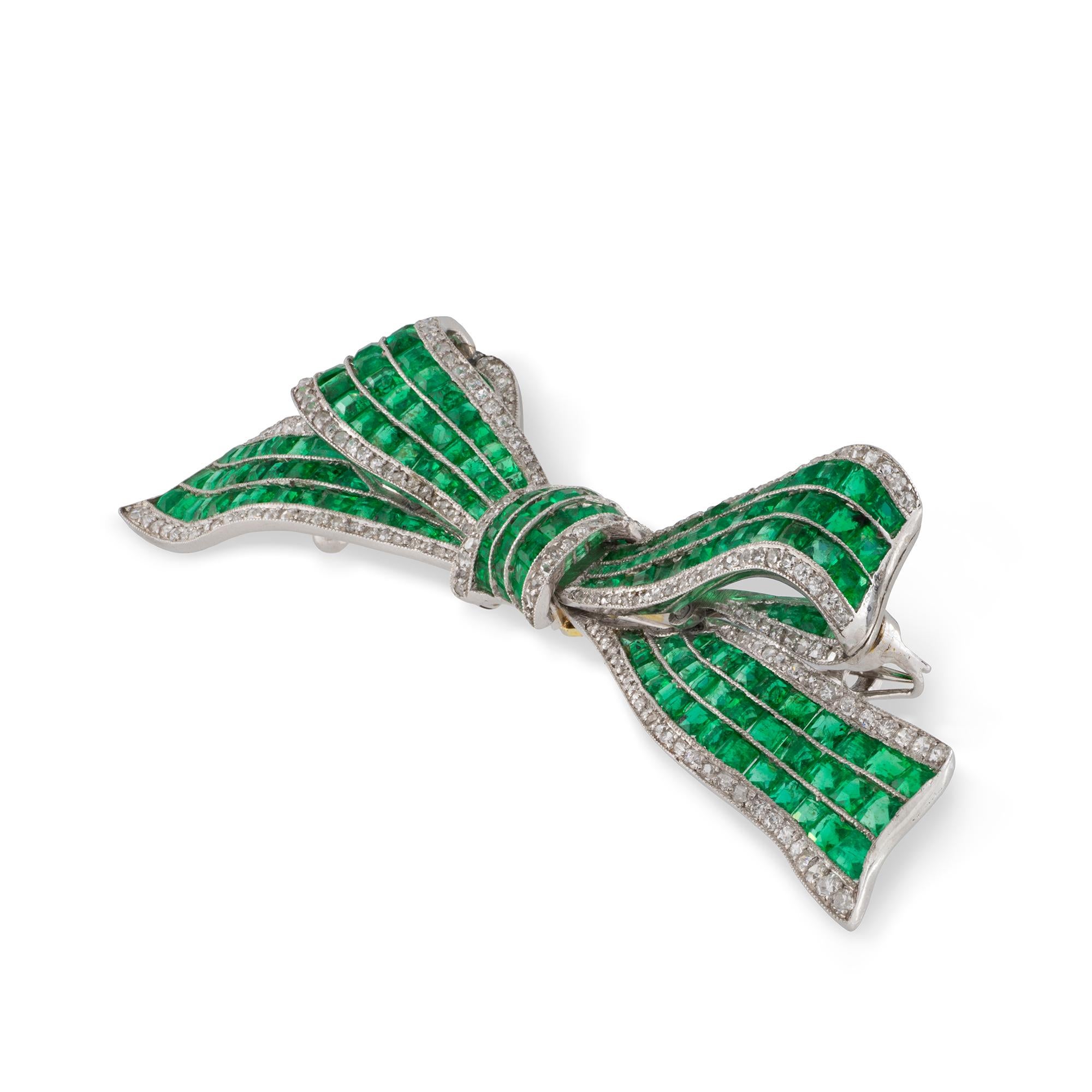 An Art Deco emerald and diamond bow brooch, the brooch in the form of a ribbon bow, set with central triple-row of calibre-cut emeralds between two rows of swiss-cut diamonds, all set in platinum mount and brooch fitting, circa 1920, measuring