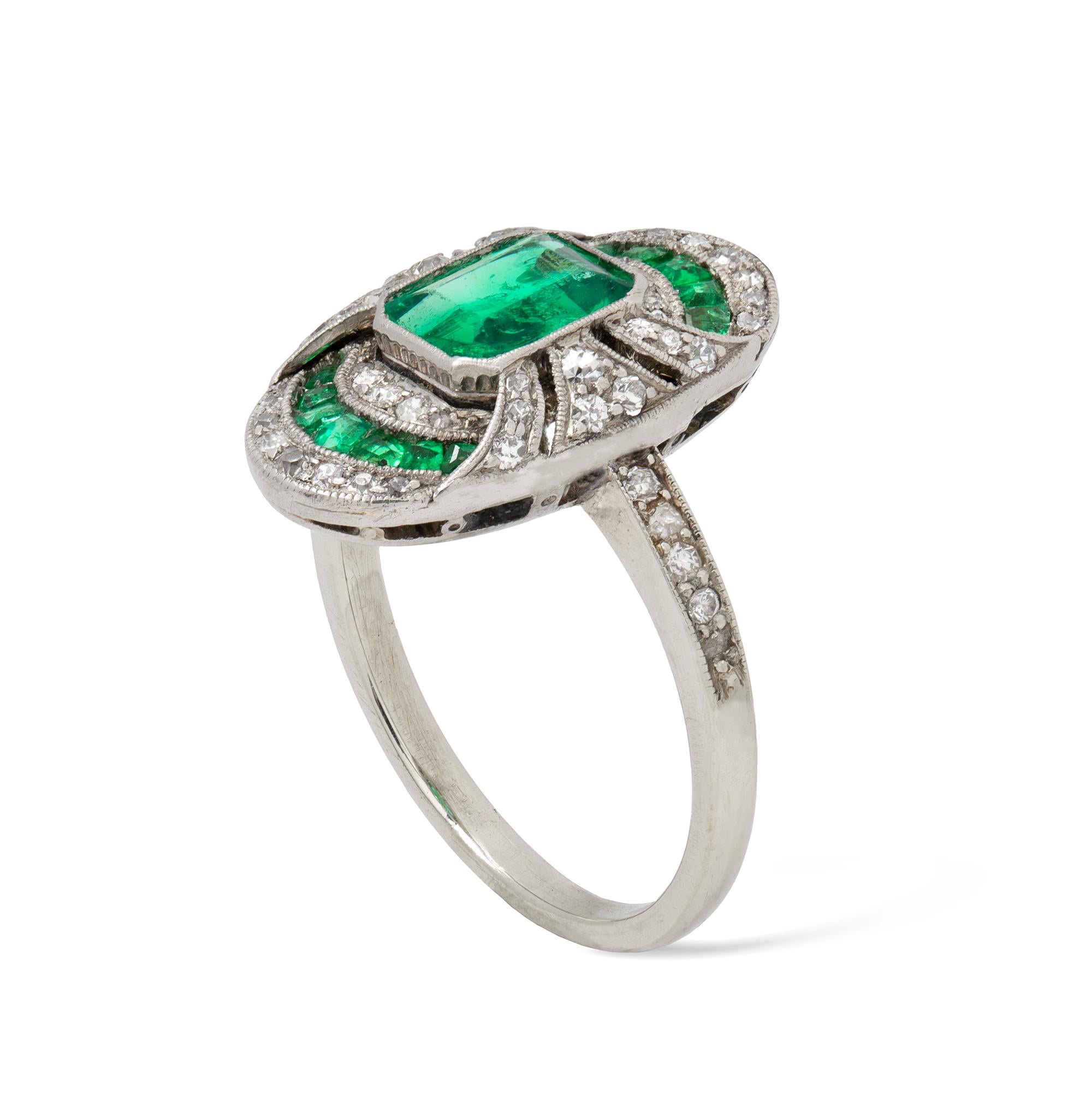 An Art Deco emerald and diamond ring, the octagonal-cut emerald with estimated weight 0.7 carats rub-over millegrain set to the centre of an oval plaque, set with old brilliant-cut diamonds and calibre-cut emeralds, all set in platinum, circa 1920,
