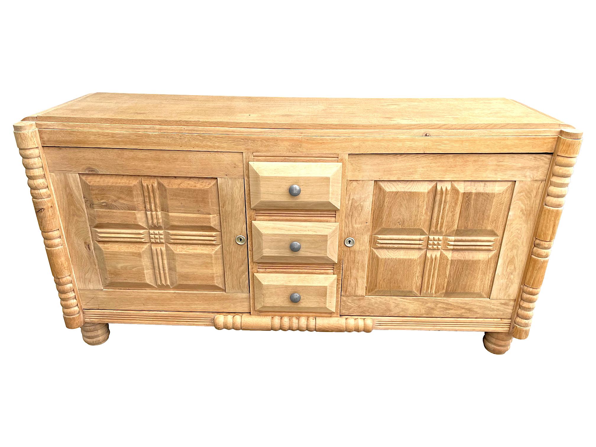An 1940s Art Deco natural oak sideboard by Gaston Poisson with geometric design on each door either side of three central drawers. The oak is a lovely natural bleached colour with brass escutcheons and orignal key with carved oak corners and central