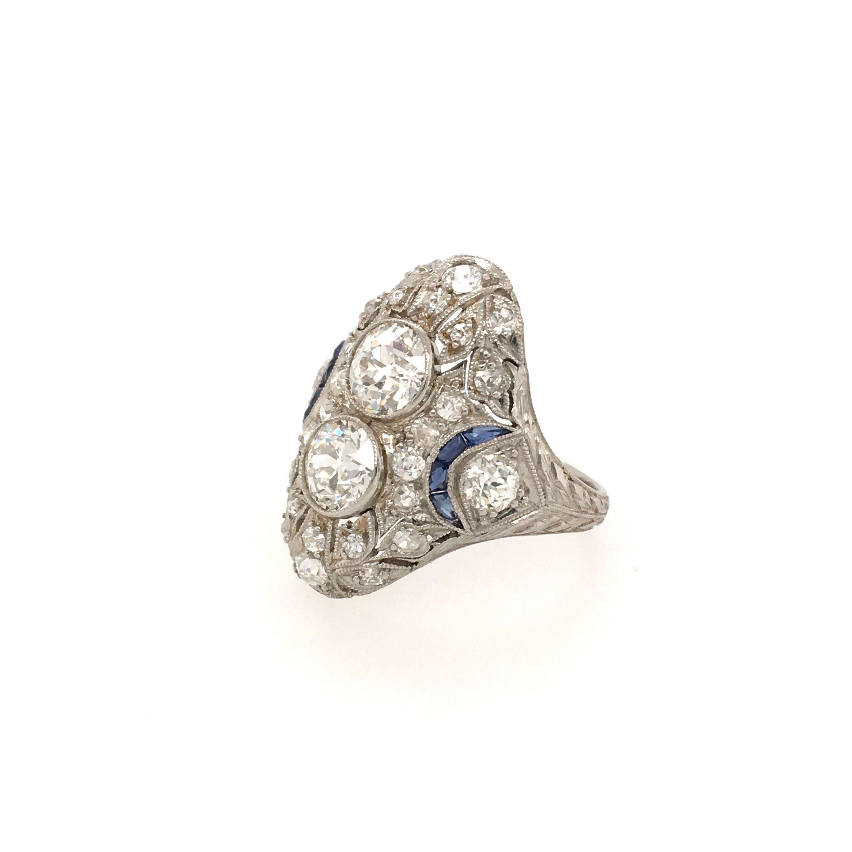 An Art Deco platinum, diamond and sapphire ring. Circa 1925. Designed as an oval pave set diamond plaque, centering two (2) old European cut diamonds, each weighing approximately 0.75 carats, enhanced by two (2) smaller old European cut diamonds and