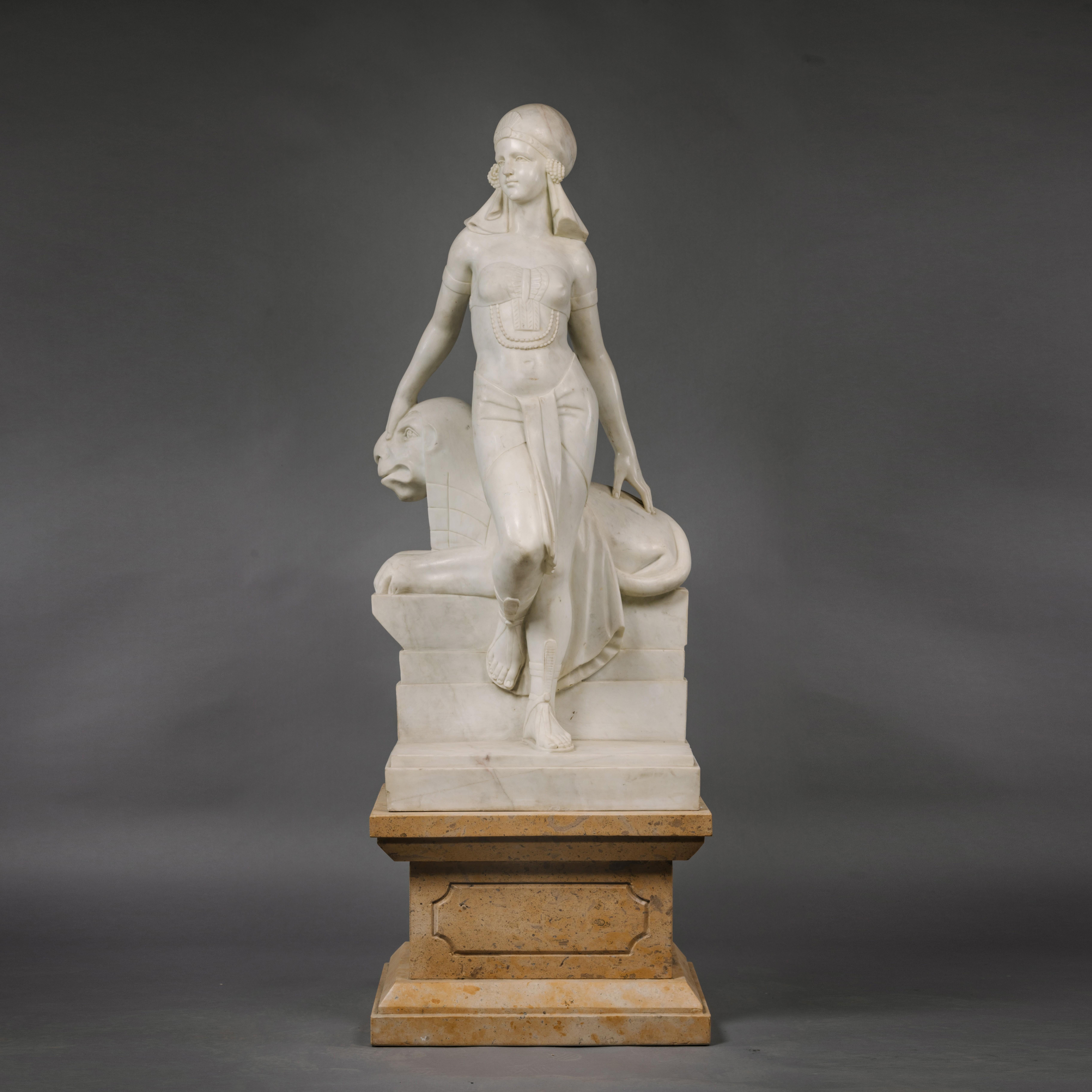 An Art Deco statuary marble figure of Cleopatra.

The half-lifesize standing figure posed with her right foot raised and looking to dexta, in front of a recumbent sphinx raised on a stepped entablature. On a Siena marble pedestal carved with